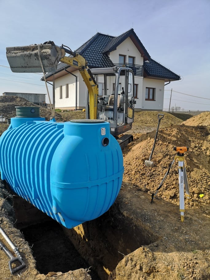 Niebieski zbiornik na szambo częściowo umieszczony w wykopie, z koparką w tle i niwelatorem na trójnogu. Widoczny dom jednorodzinny.
