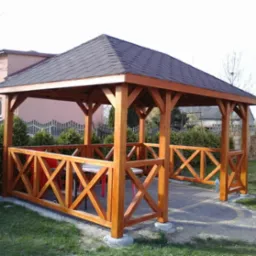 Drewniana altana z ciemnym dachem, otoczona zielenią i kostką brukową. Widoczne detale konstrukcji oraz krzyżowe zdobienia barierek. Stolik wewnątrz.