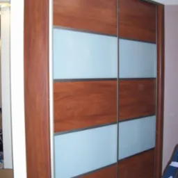 Wysoka szafa wnękowa z przesuwnymi drzwiami, wykonana z drewna o ciepłym odcieniu, urozmaicona poziomymi pasami z matowego szkła. Widoczny detal wykończenia.