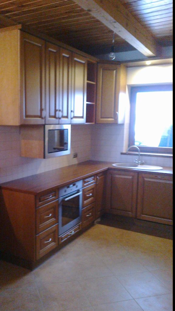 Drewniana kuchnia z frezowanymi frontami, zabudowaną kuchenką i mikrofalówką. Widoczny fragment sufitu z drewnianymi belkami i okno z naturalnym światłem.