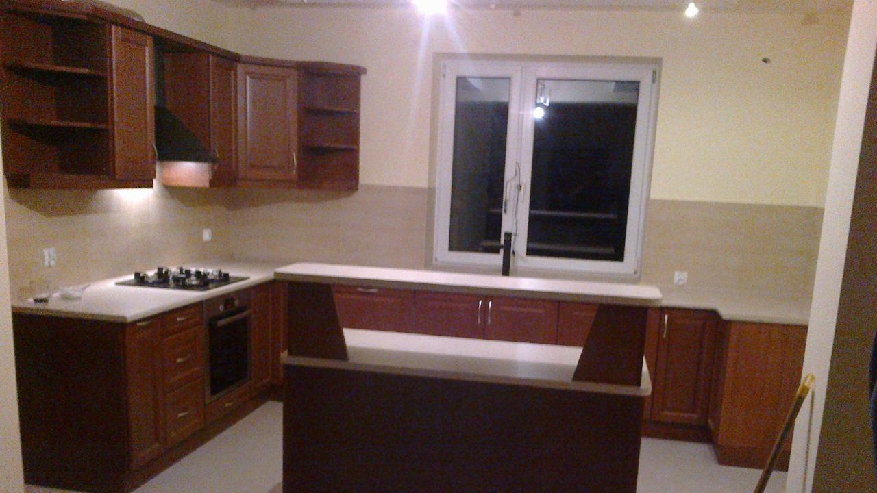 Kuchnia z drewnianymi szafkami i wyspą na środku, oświetlona lampami sufitowymi. Widoczne okno i sprzęt AGD. Blaty w jasnym odcieniu.