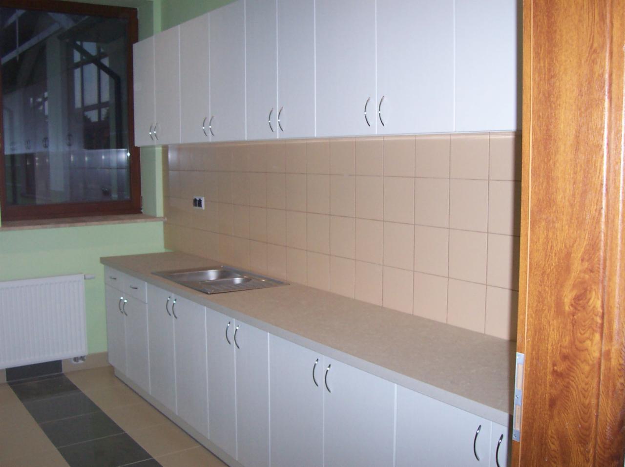 Jasna kuchnia z białymi szafkami, beżowym blatem i płytkami. Widoczny zlew, okno i grzejnik. Drzwi z prawej strony kadru. Ujęcie całościowe, brak detali.