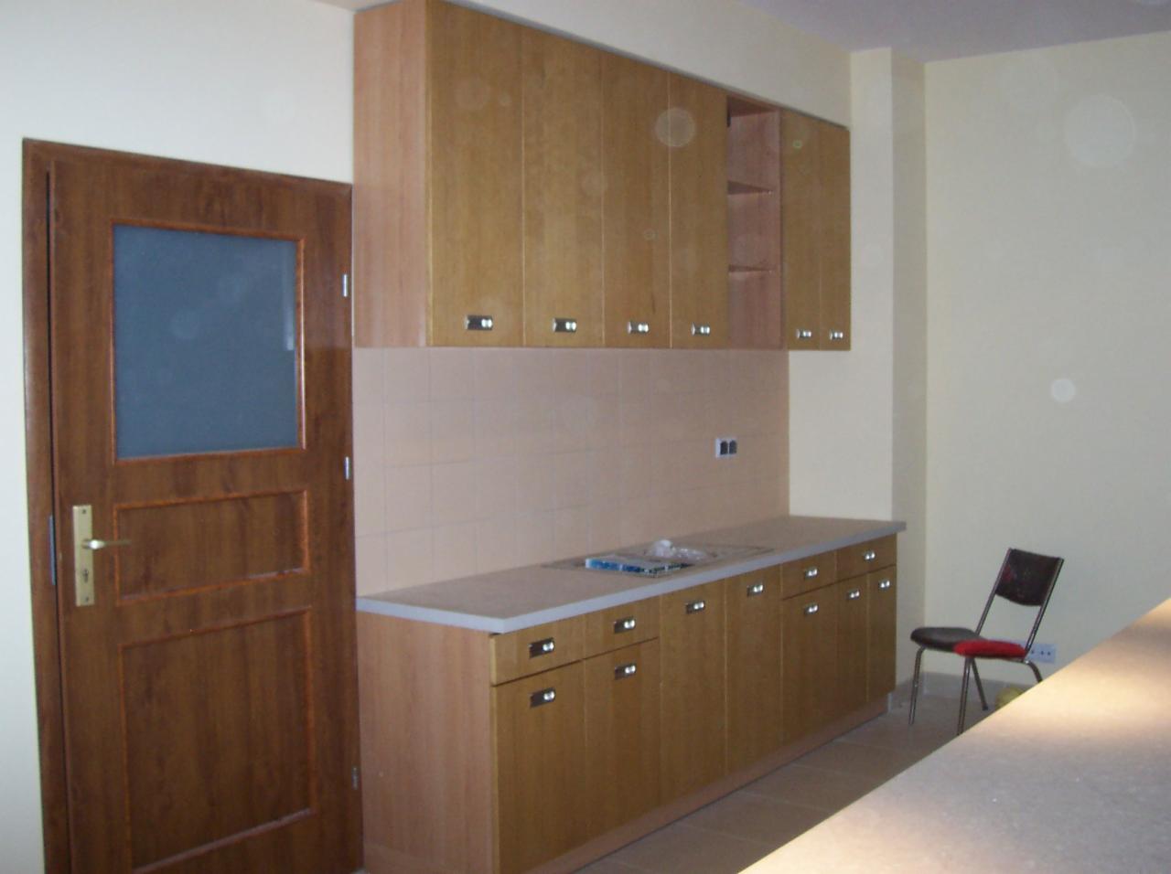 Jasna kuchnia z drewnianymi meblami: szafki wiszące i dolne, beżowe płytki nad blatem. Obok brązowe drzwi z szybą i krzesło. Widok na fragment blatu.