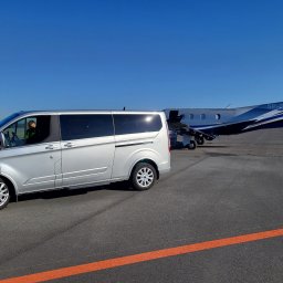 Dominik Szymocha - Srebrny van na lotnisku w Myślachowicach, z widocznym pilotem w kabinie i prywatnym samolotem w tle, sugerujący ekskluzywny transport.