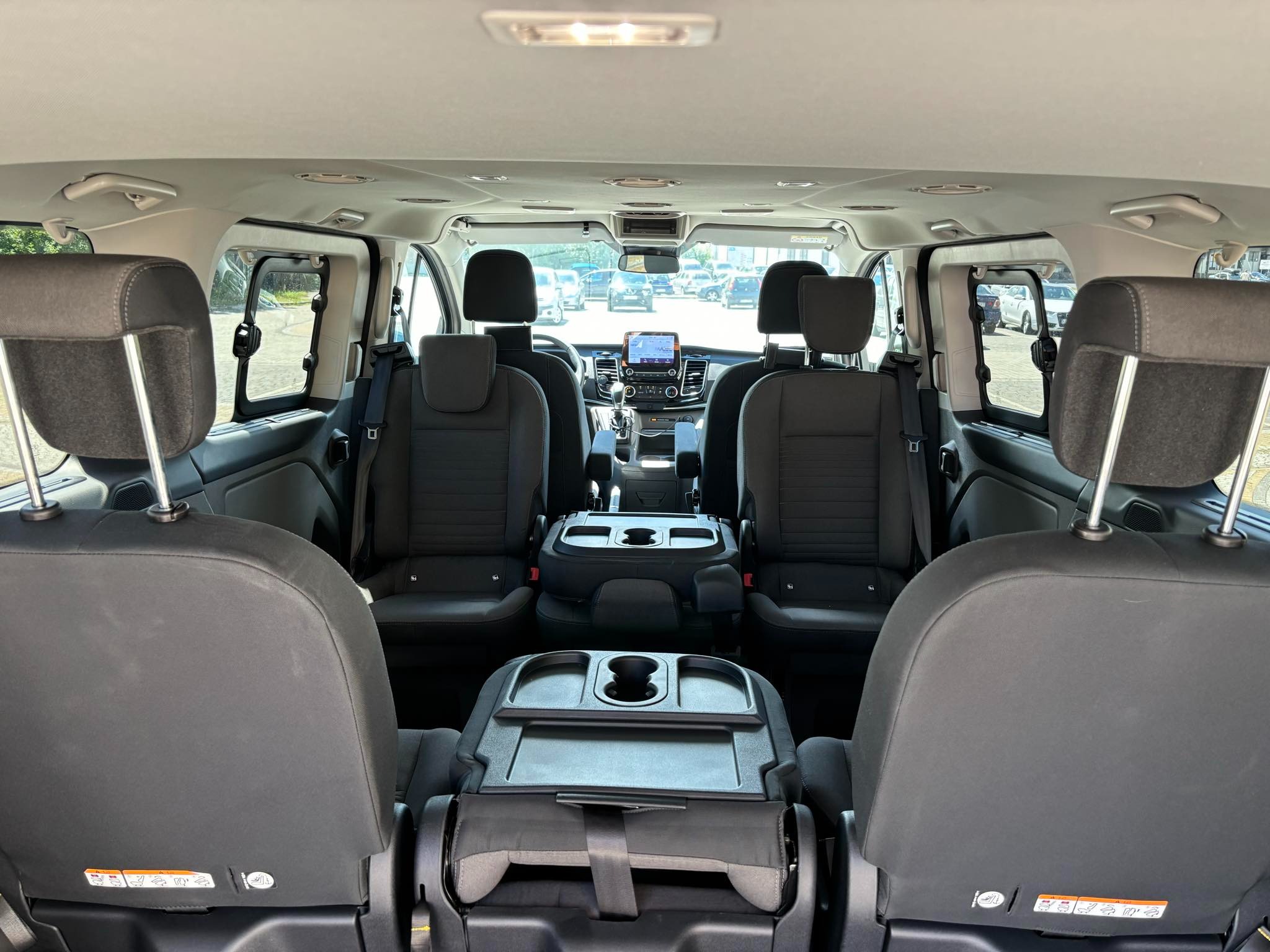 Wnętrze busa pasażerskiego z czarnymi fotelami, widok z tyłu na przód pojazdu, z widocznymi zagłówkami, pasami bezpieczeństwa i stolikami między siedzeniami.