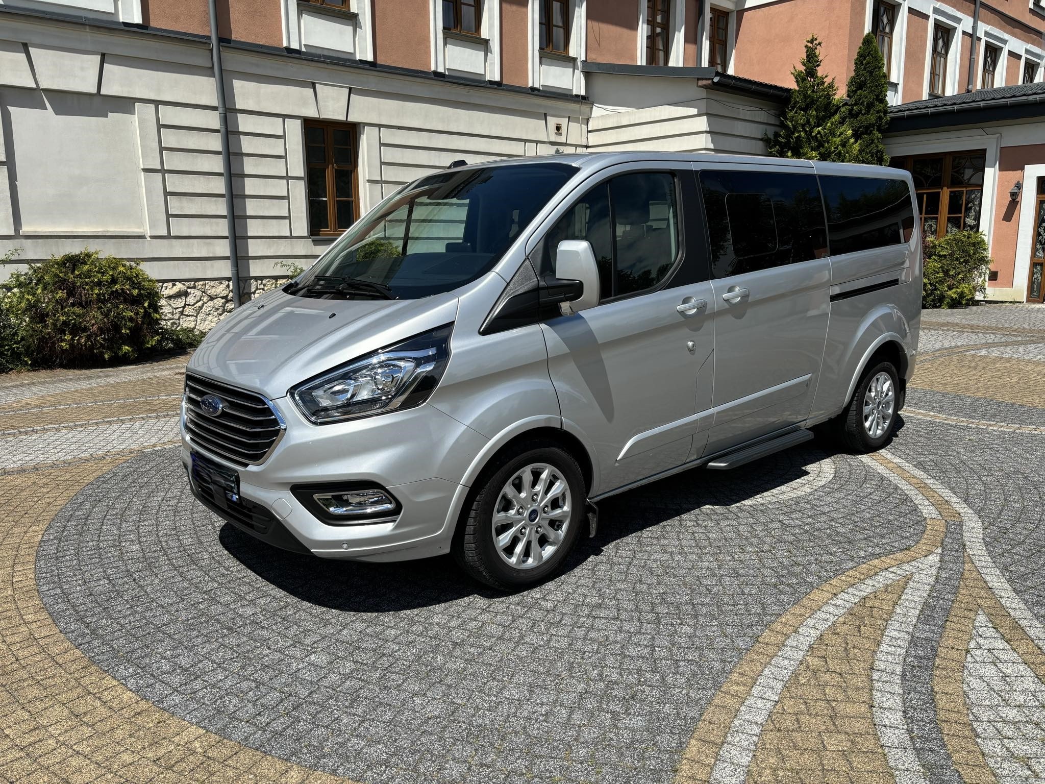 Srebrny bus Ford Transit Custom z wysuwanym stopniem, zaparkowany na ozdobnej kostce brukowej przed budynkiem.