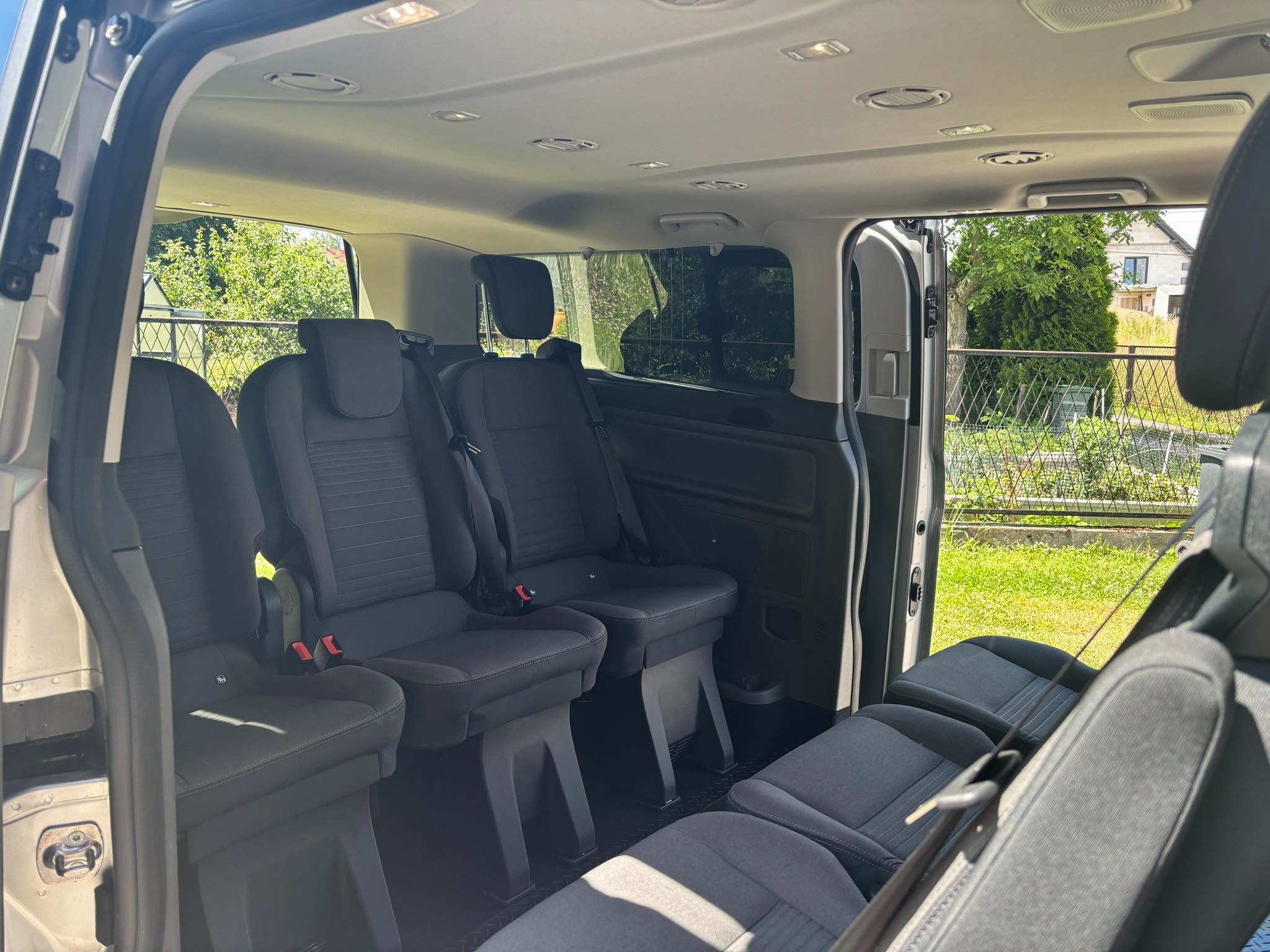 Wnętrze przestronnego busa z trzema rzędami szarych foteli, widok z otwartych drzwi na zielony ogród w tle.