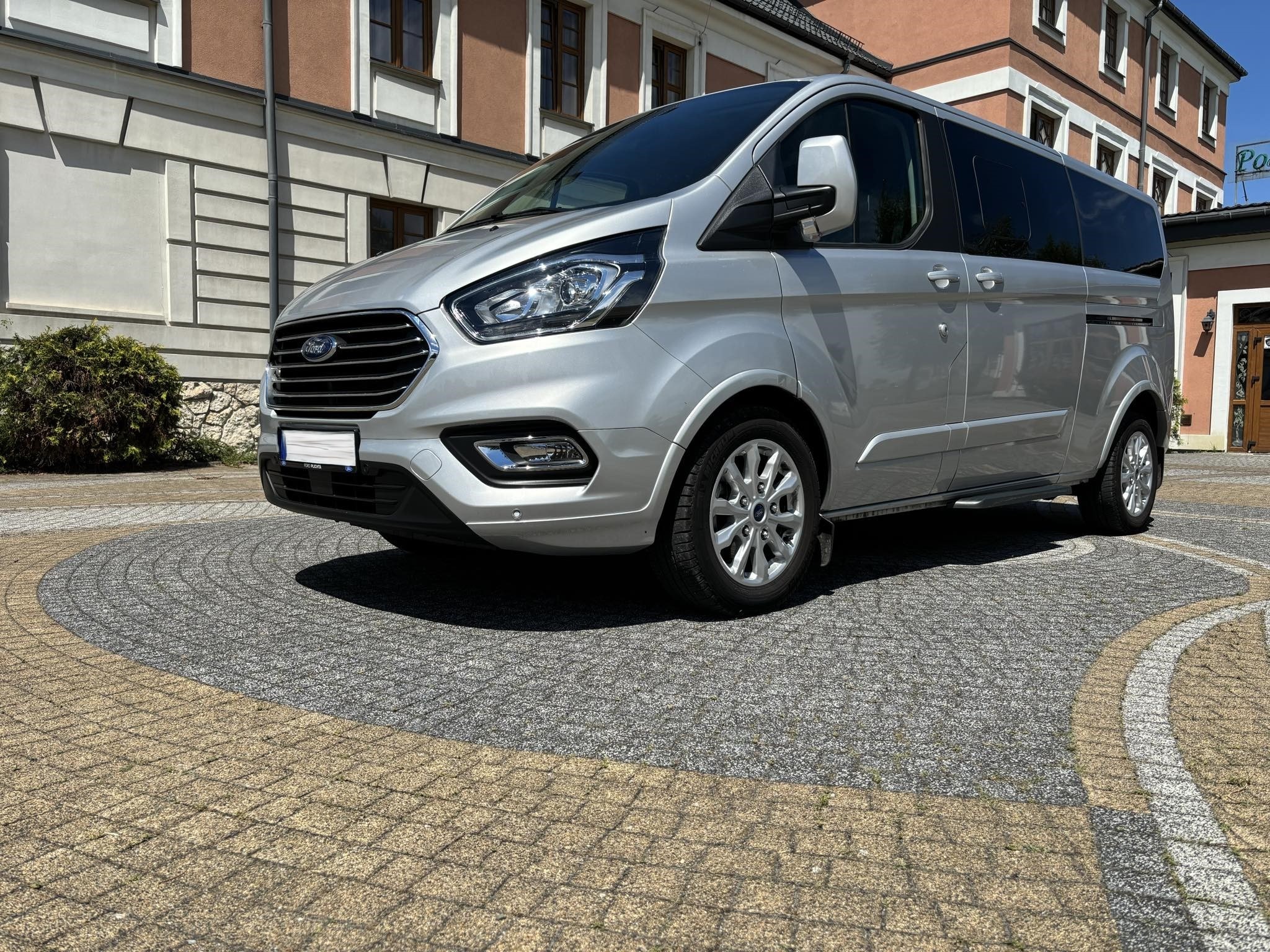 Srebrny Ford Transit Custom na ozdobnej kostce brukowej przed budynkiem w klasycznym stylu.