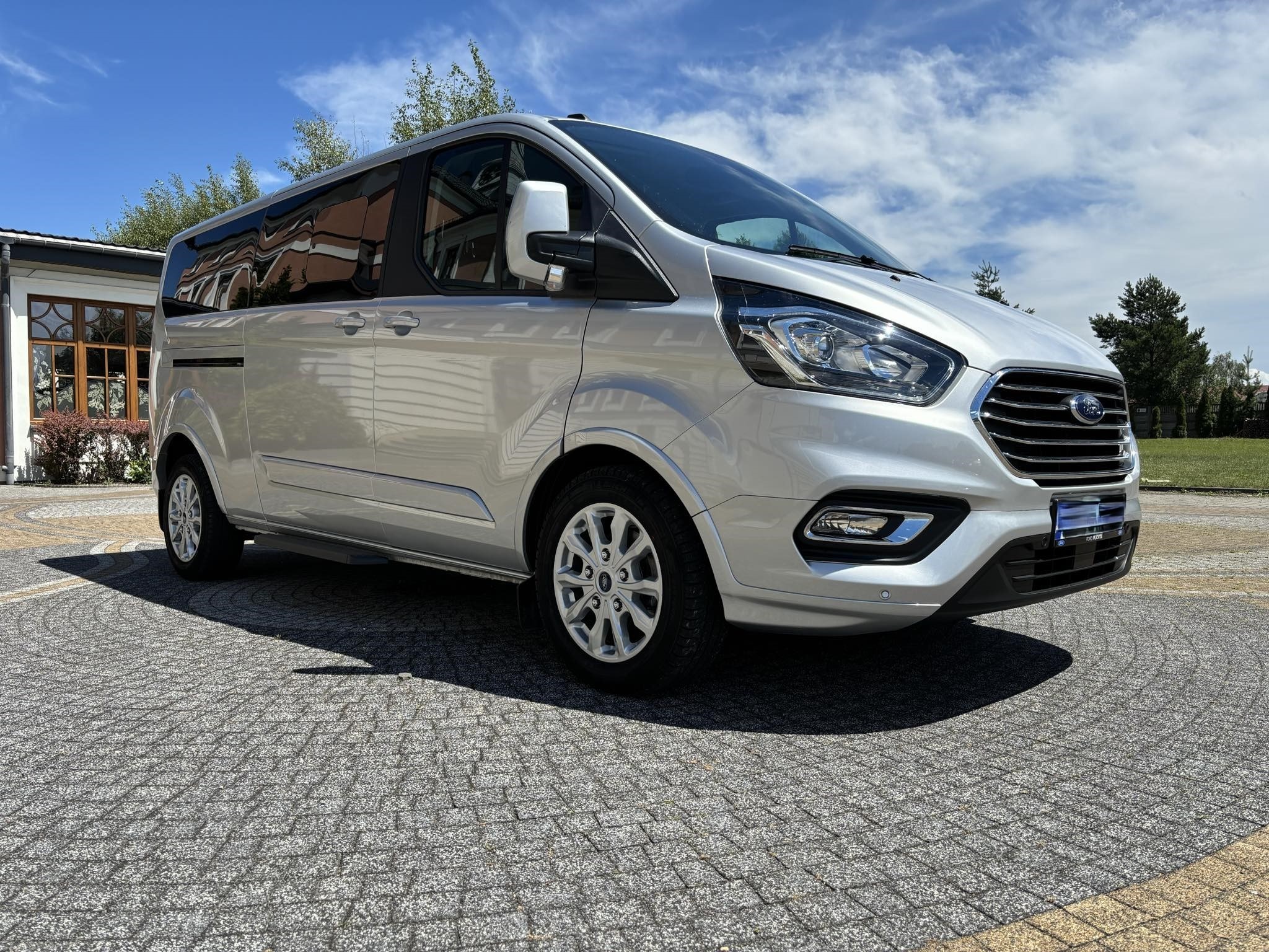 Srebrny Ford Transit Custom Kombi na brukowanym podjeździe w słoneczny dzień.