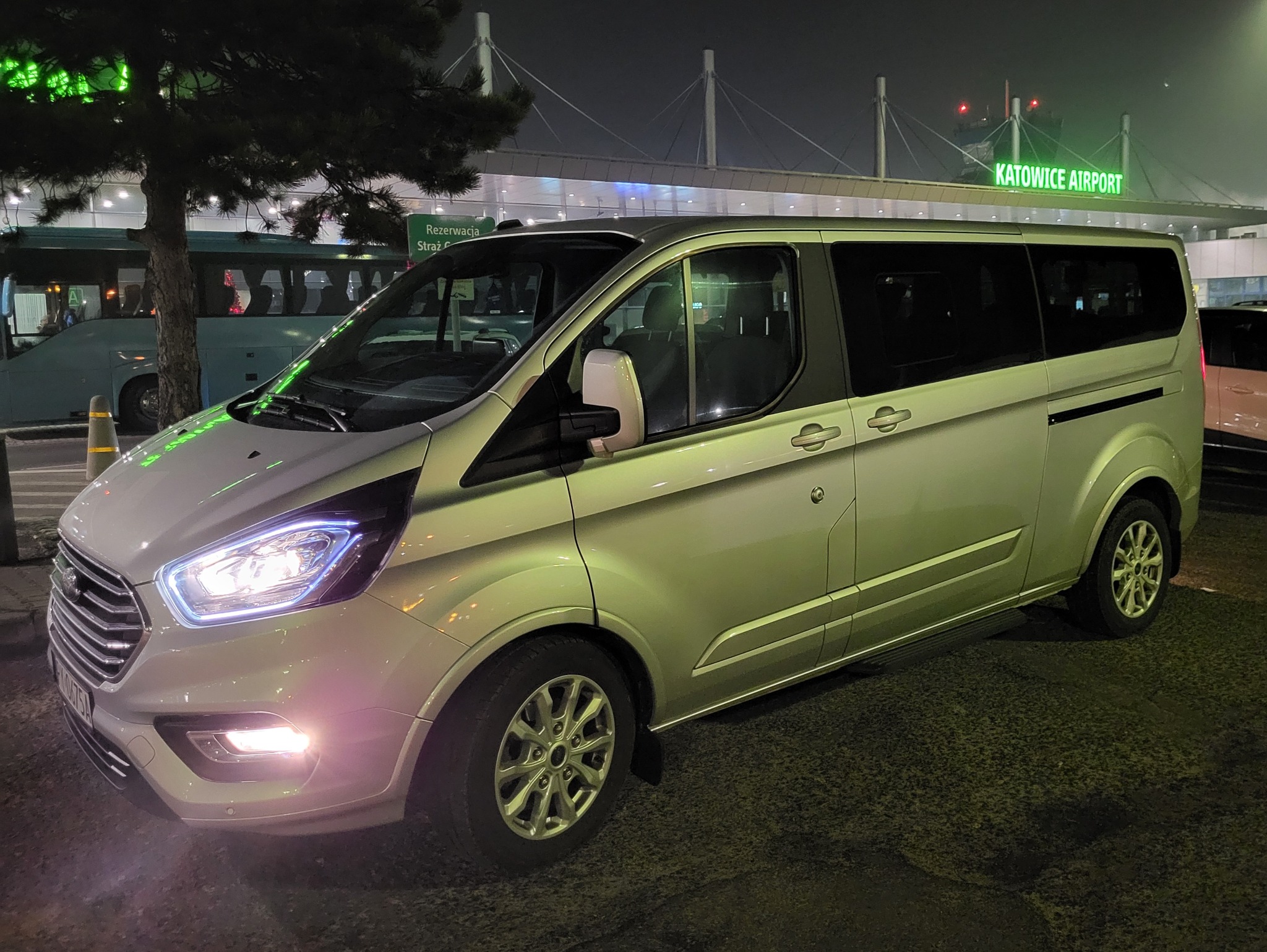 Srebrny van Ford Transit Custom z włączonymi światłami postojowymi, zaparkowany nocą przed budynkiem lotniska Katowice Airport.