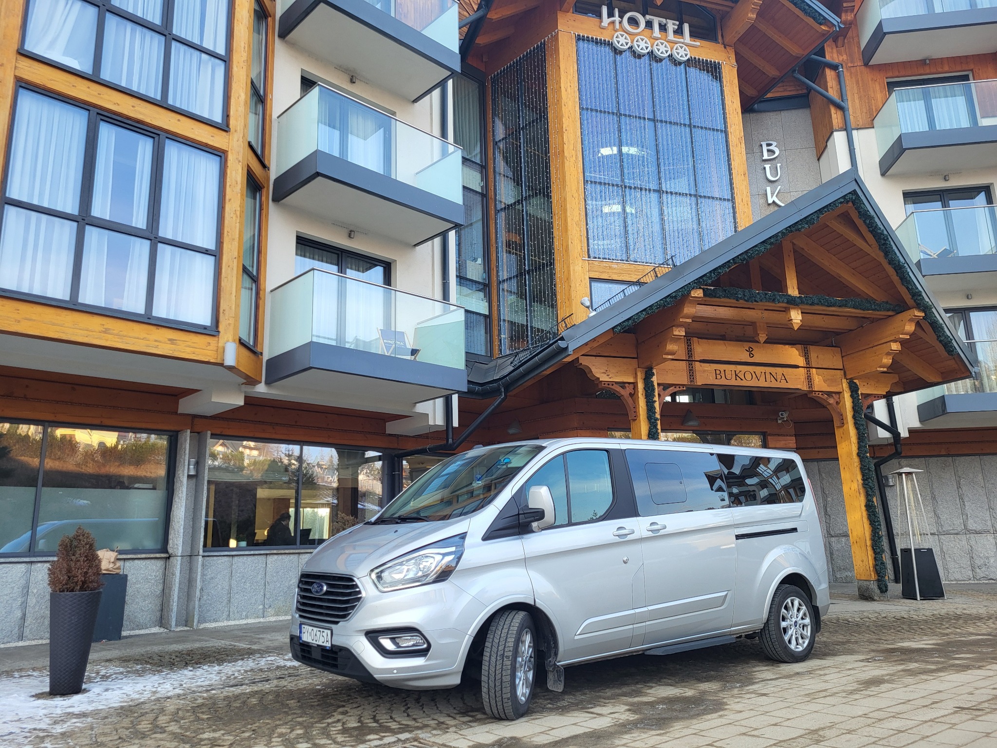Srebrny van Ford Transit Custom zaparkowany przed hotelem Bukovina w słoneczny dzień, widoczne detale karoserii i otoczenia.