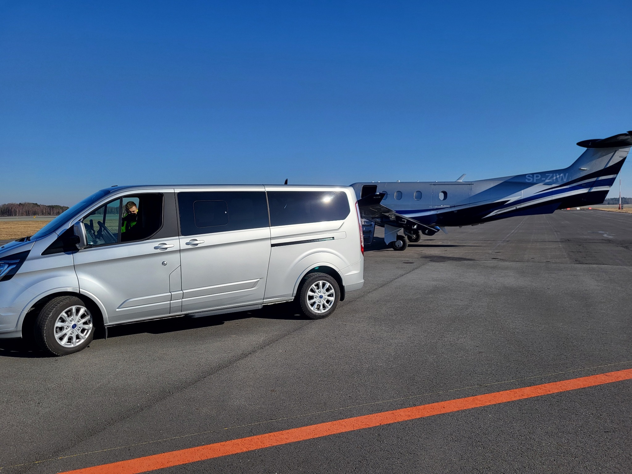 Srebrny van na lotnisku w Myślachowicach, z widocznym pilotem w kabinie i prywatnym samolotem w tle, sugerujący ekskluzywny transport.