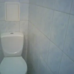 Biała toaleta z widoczną klapą rewizyjną na ścianie, wykończonej jasnoniebieskimi płytkami ceramicznymi.