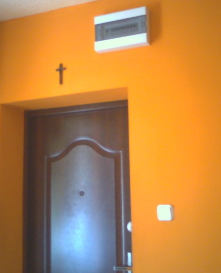 Pomarańczowa ściana z brązowymi drzwiami, skrzynką elektryczną i krzyżykiem nad futryną.