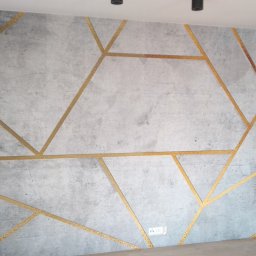 Kamil Furmanek - Ściana wykończona szarą tapetą imitującą beton z geometrycznym wzorem złotych linii, widoczne gniazdka elektryczne.