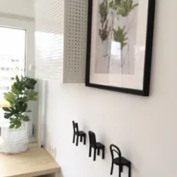 Jasne wnętrze z wiszącą szafką z perforowanej blachy, oprawionym obrazem z motywem roślinnym i trzema małymi, czarnymi wieszakami w kształcie krzeseł na białej ścianie, obok blatu z rośliną...