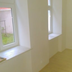 Kamil Furmanek - Pomalowane na biało wnętrze z dwoma oknami, jasną podłogą i gładkimi ścianami, widoczne gniazdko elektryczne.