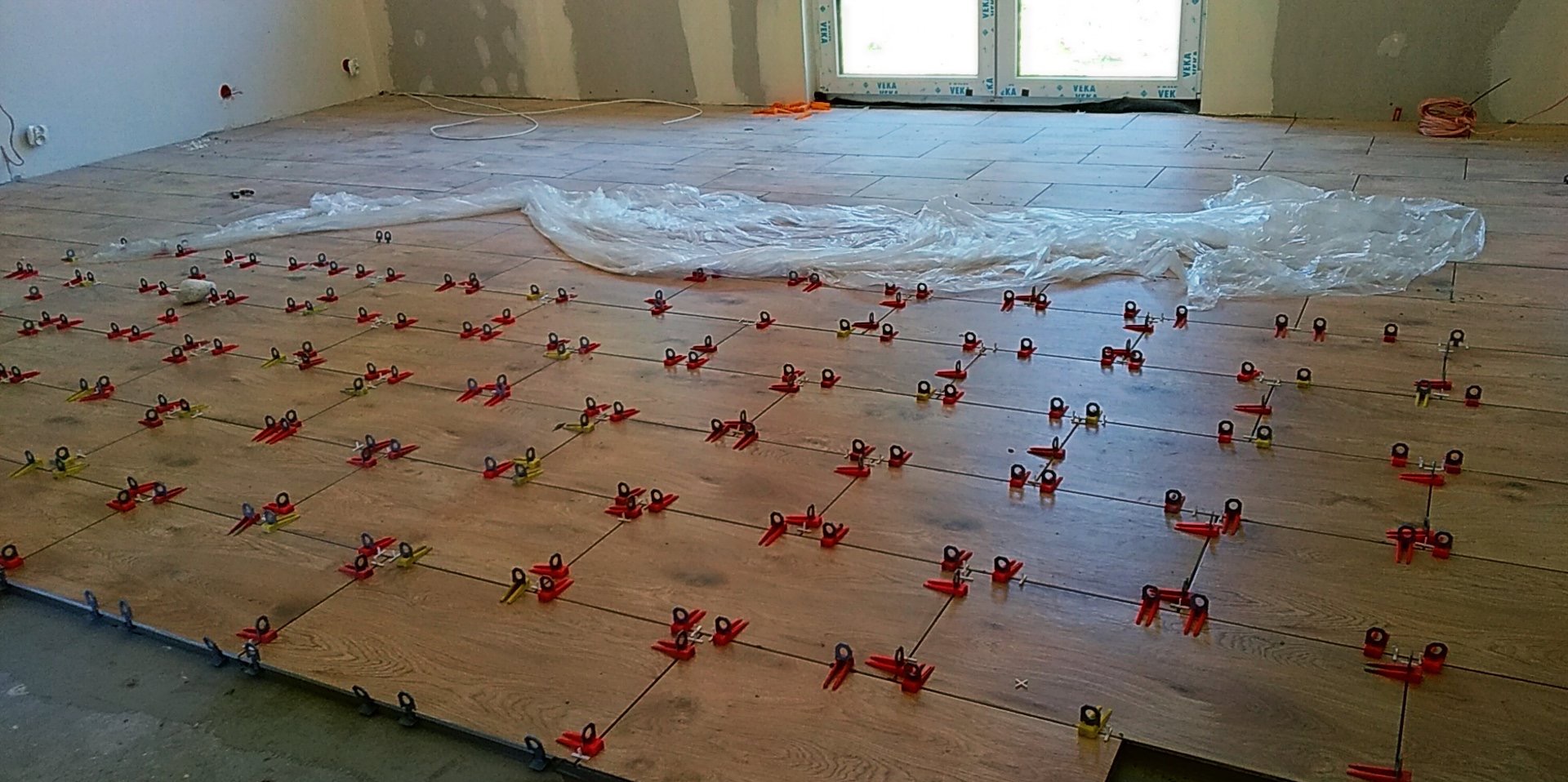 Układanie płytek podłogowych imitujących drewno, z użyciem systemu poziomowania z czerwono-żółtymi klipsami, na podłodze w trakcie prac wykończeniowych w jasnym pomieszczeniu.