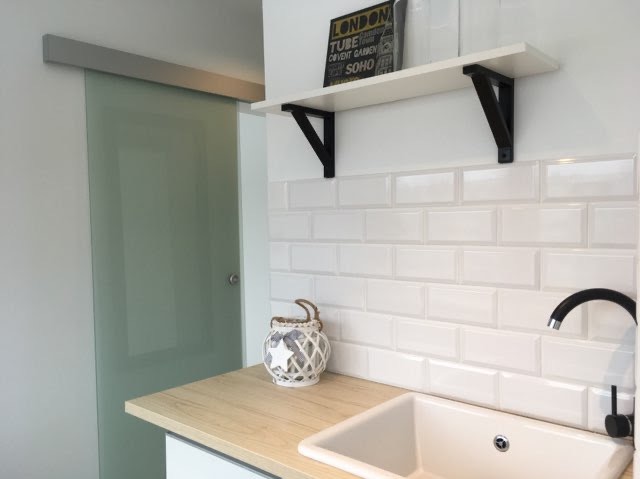 Biała umywalka w łazience z białymi płytkami typu metro, drewniany blat, dekoracyjny lampion i szklane drzwi przesuwne.