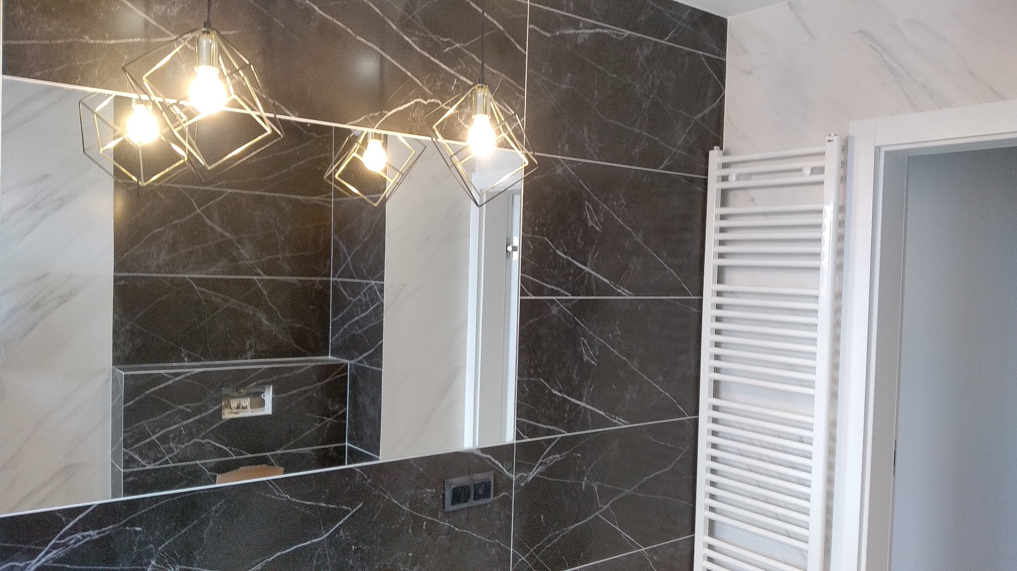 Wykończona łazienka z czarnymi płytkami imitującymi marmur, ozdobnym grzejnikiem drabinkowym, lustrem i geometrycznymi lampami wiszącymi
