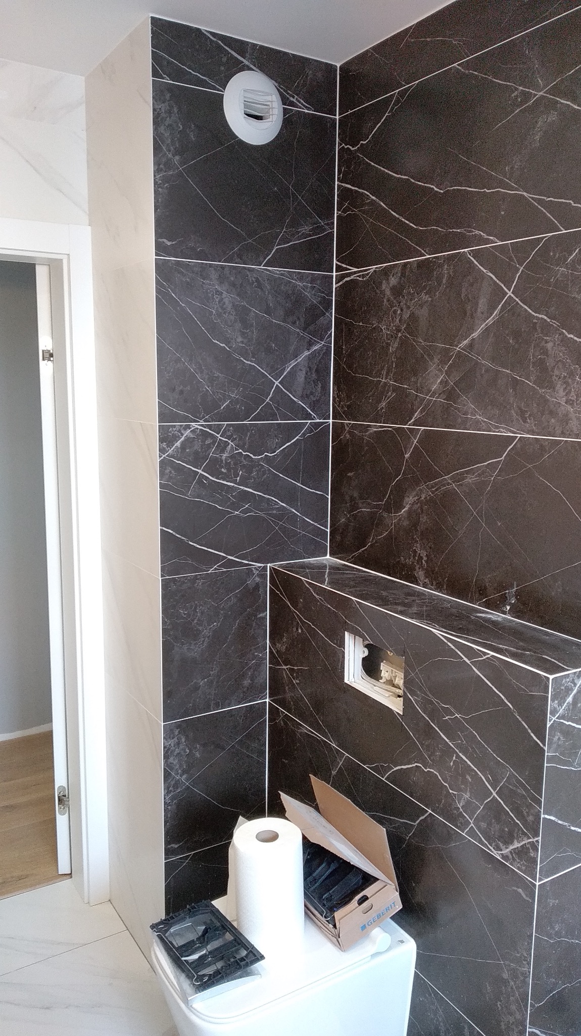 Częściowo wykończona łazienka z czarnymi płytkami imitującymi marmur z białymi żyłami, widoczny biały sedes z rolką papieru toaletowego i otwartym pudełkiem z elementami montażowymi Geberit, biały...