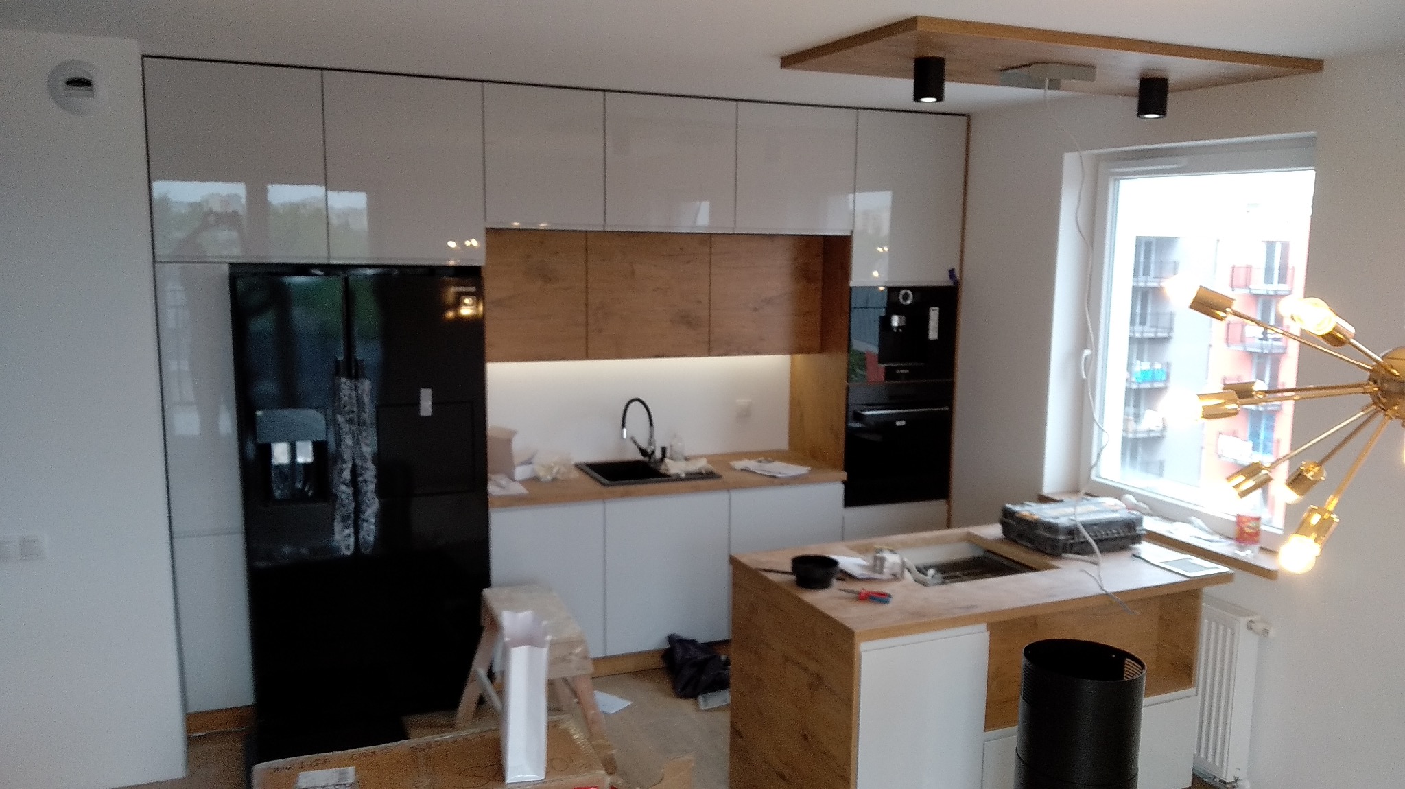 Nowoczesna kuchnia z białymi szafkami, drewnianymi akcentami i czarną lodówką side-by-side, widok na fragment blatu roboczego z narzędziami sugerujący prace wykończeniowe.