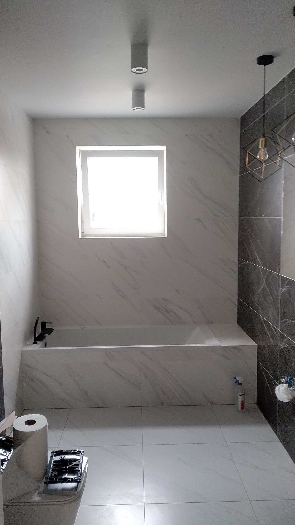 Nowoczesna łazienka z wanną obłożoną białymi płytkami imitującymi marmur, kontrastującymi z ciemnoszarymi płytkami na ścianie obok, widoczne białe podwieszane lampy sufitowe i dekoracyjna lampa...