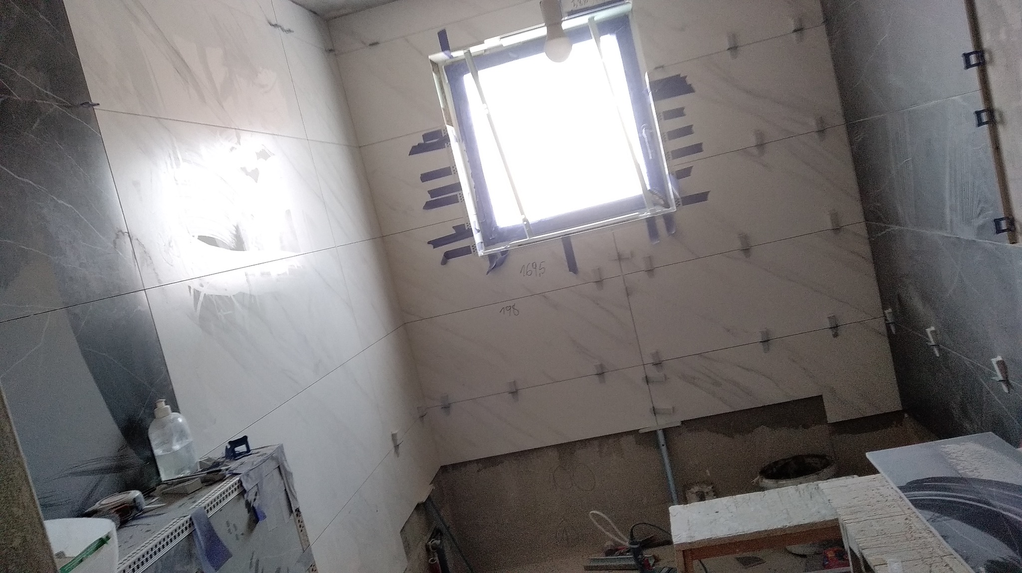 W trakcie układania płytek wielkoformatowych w łazience, widoczne krzyżyki dystansowe, poziomice laserowe i okno z taśmą malarską zabezpieczającą ramę.