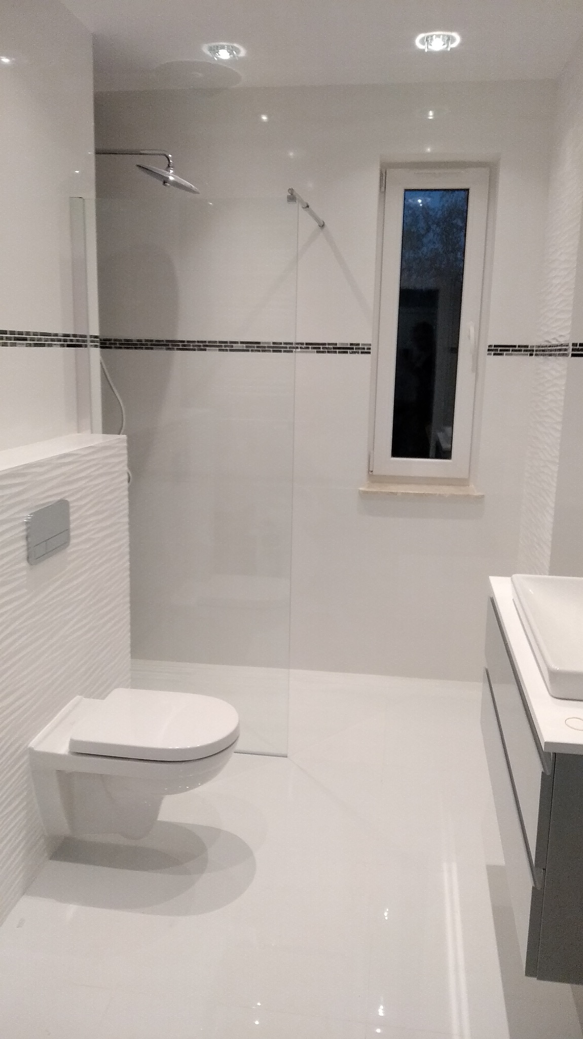 Nowoczesna, minimalistyczna łazienka z białymi płytkami, kabiną prysznicową typu walk-in, podwieszaną toaletą z fakturą na ścianie, szarym blatem umywalkowym i oknem z widokiem na zewnątrz.