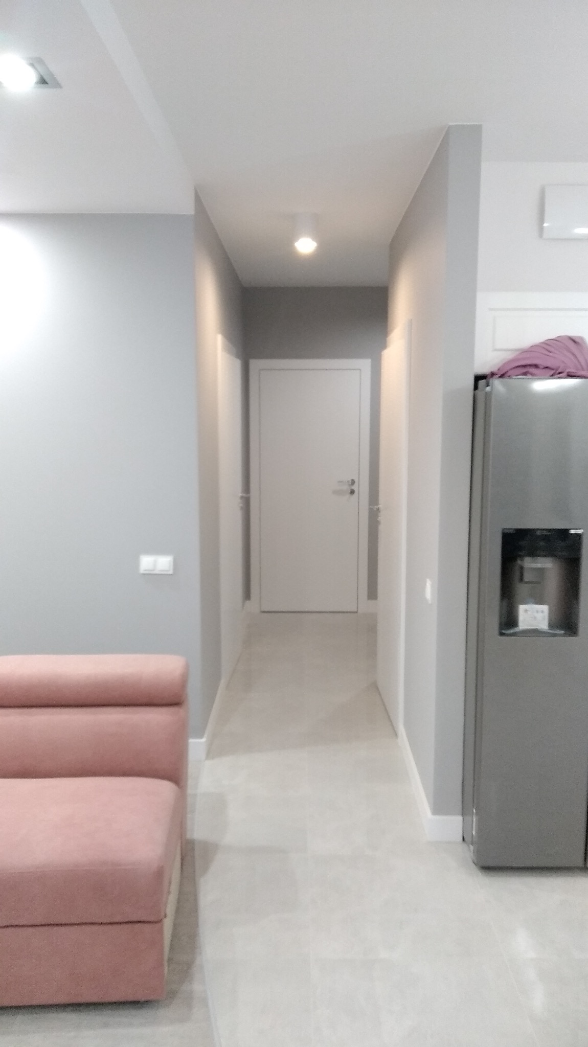 Jasne, odnowione wnętrze mieszkania z szarą kolorystyką ścian i podłogi, widoczny fragment różowej sofy, białe drzwi na wprost oraz nowoczesna lodówka z dystrybutorem wody po prawej stronie.