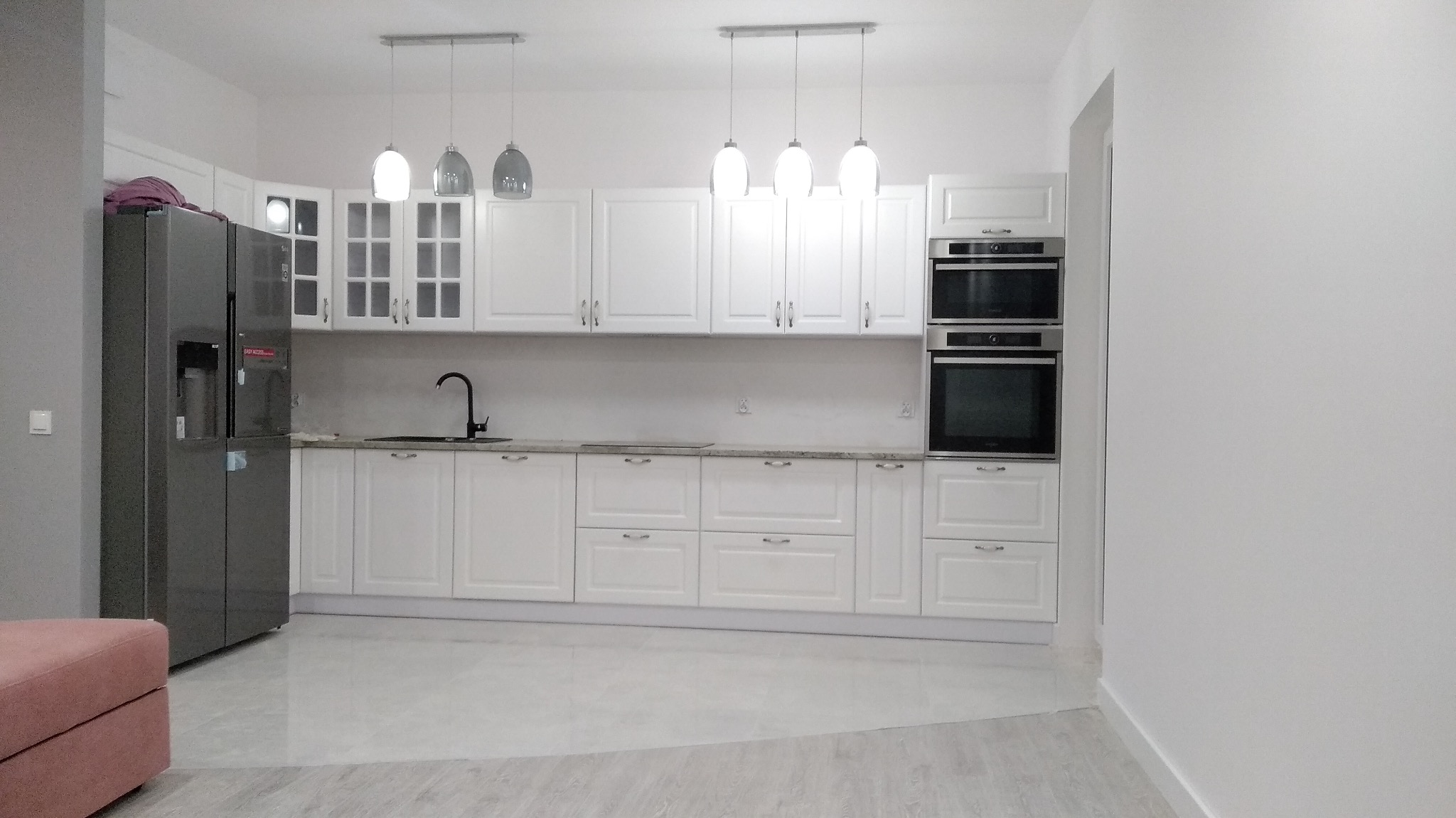 Jasna, minimalistyczna kuchnia z białymi szafkami, szarym blatem i nowoczesnym, dwudrzwiowym lodówko-zamrażarką LG, oświetlona wiszącymi lampami.