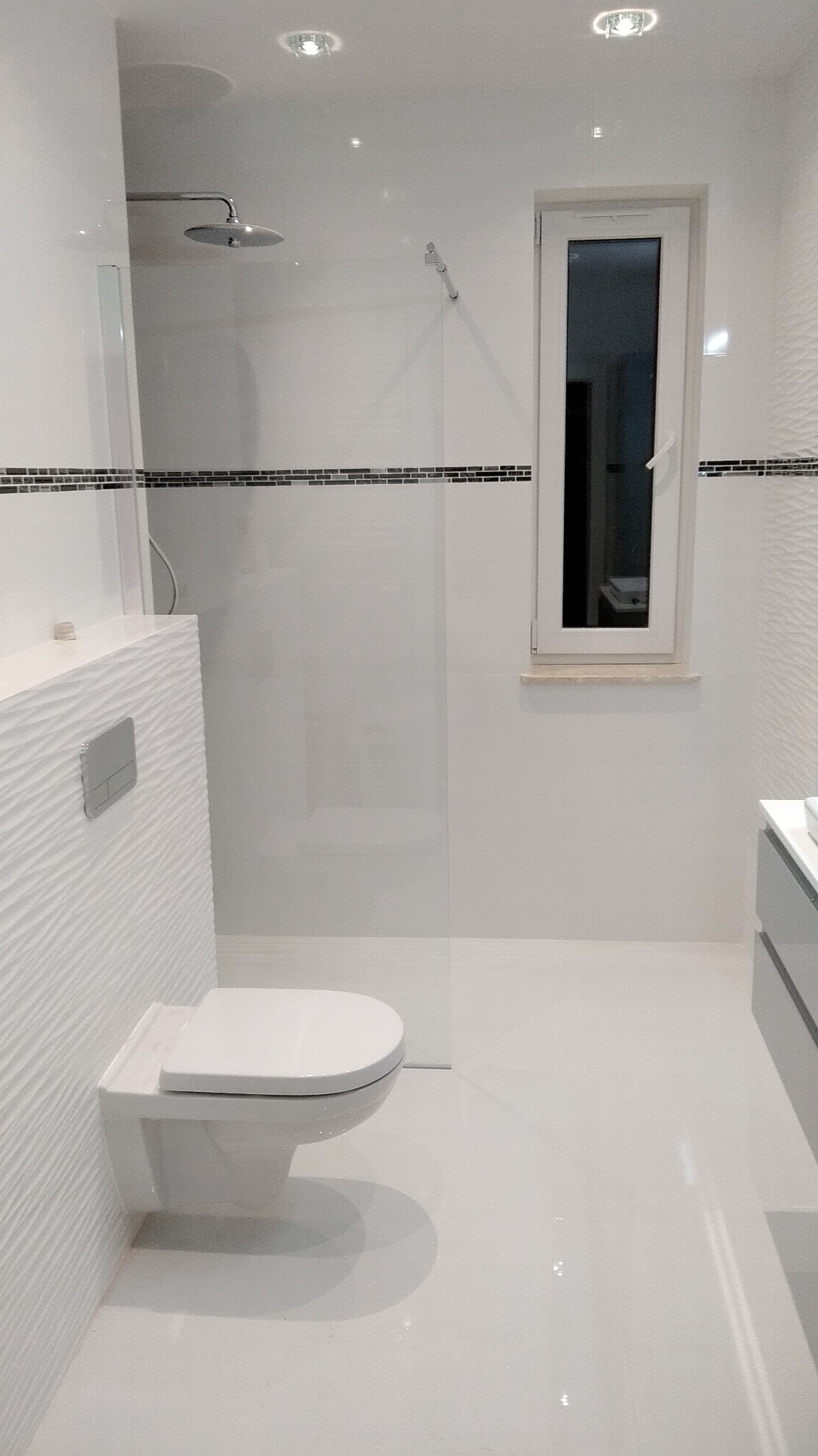 Nowoczesna, minimalistyczna łazienka z wiszącą toaletą, szklaną kabiną prysznicową typu walk-in i oknem, wykończona białymi płytkami z dekoracyjnym paskiem mozaiki.