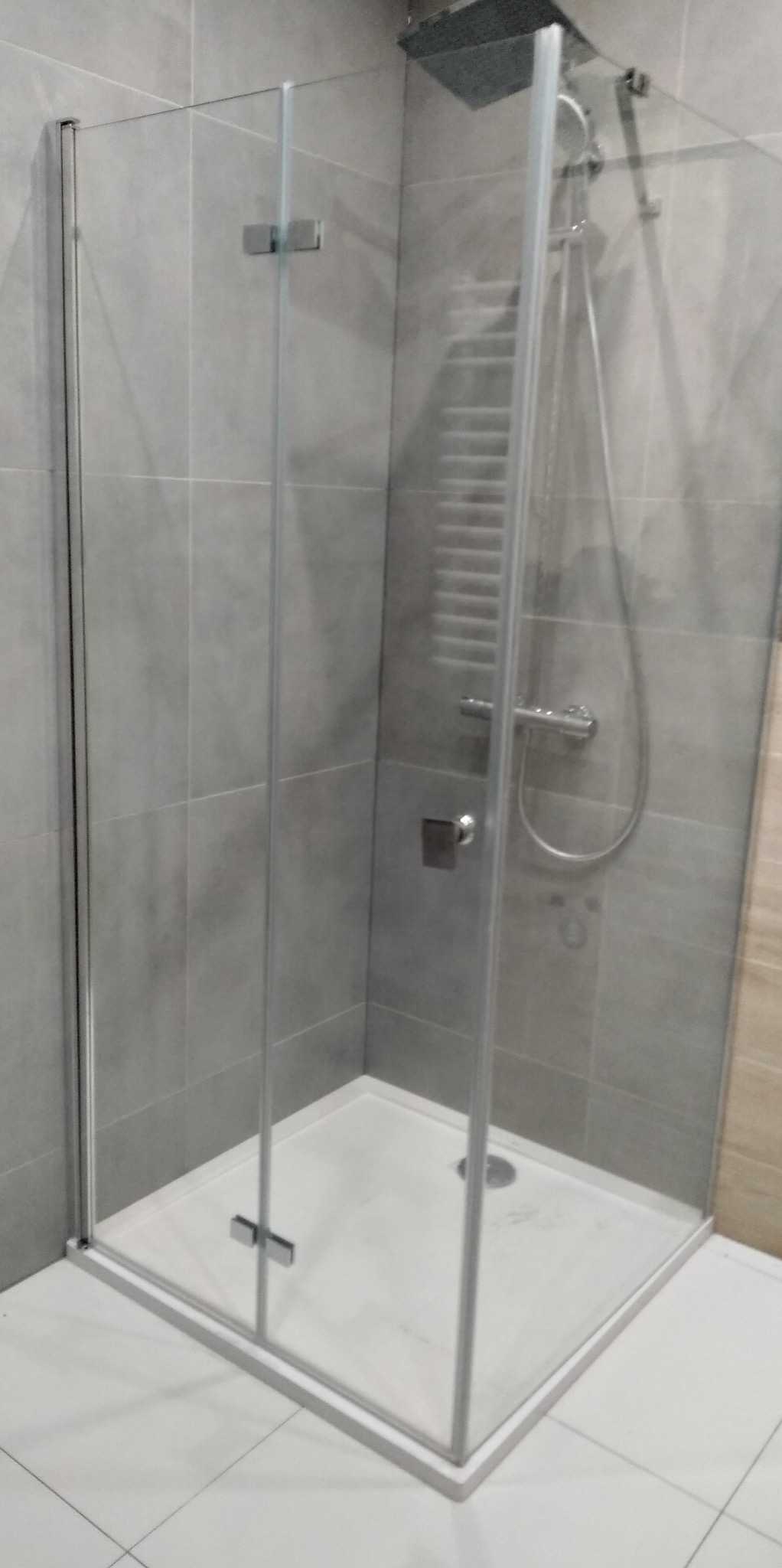 Szklana kabina prysznicowa z odpływem liniowym, szare płytki ścienne i białe podłogowe, bateria termostatyczna z deszczownicą.