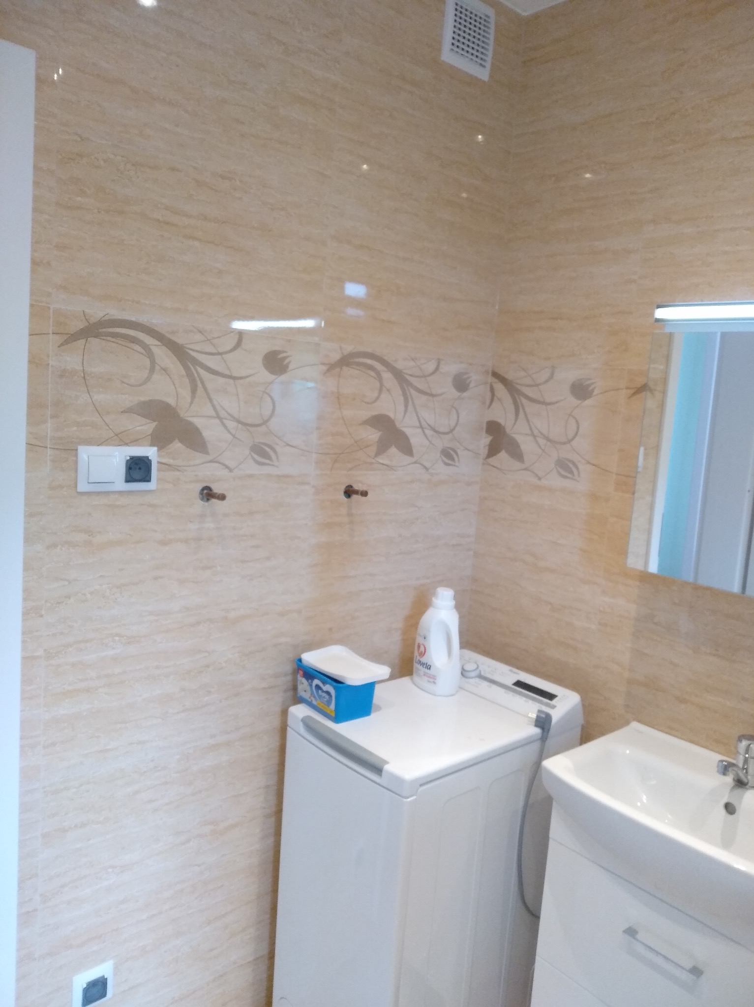 Beżowa łazienka z dekoracyjnymi płytkami, pralką, umywalką z szafką i lustrem, widoczne akcesoria łazienkowe.