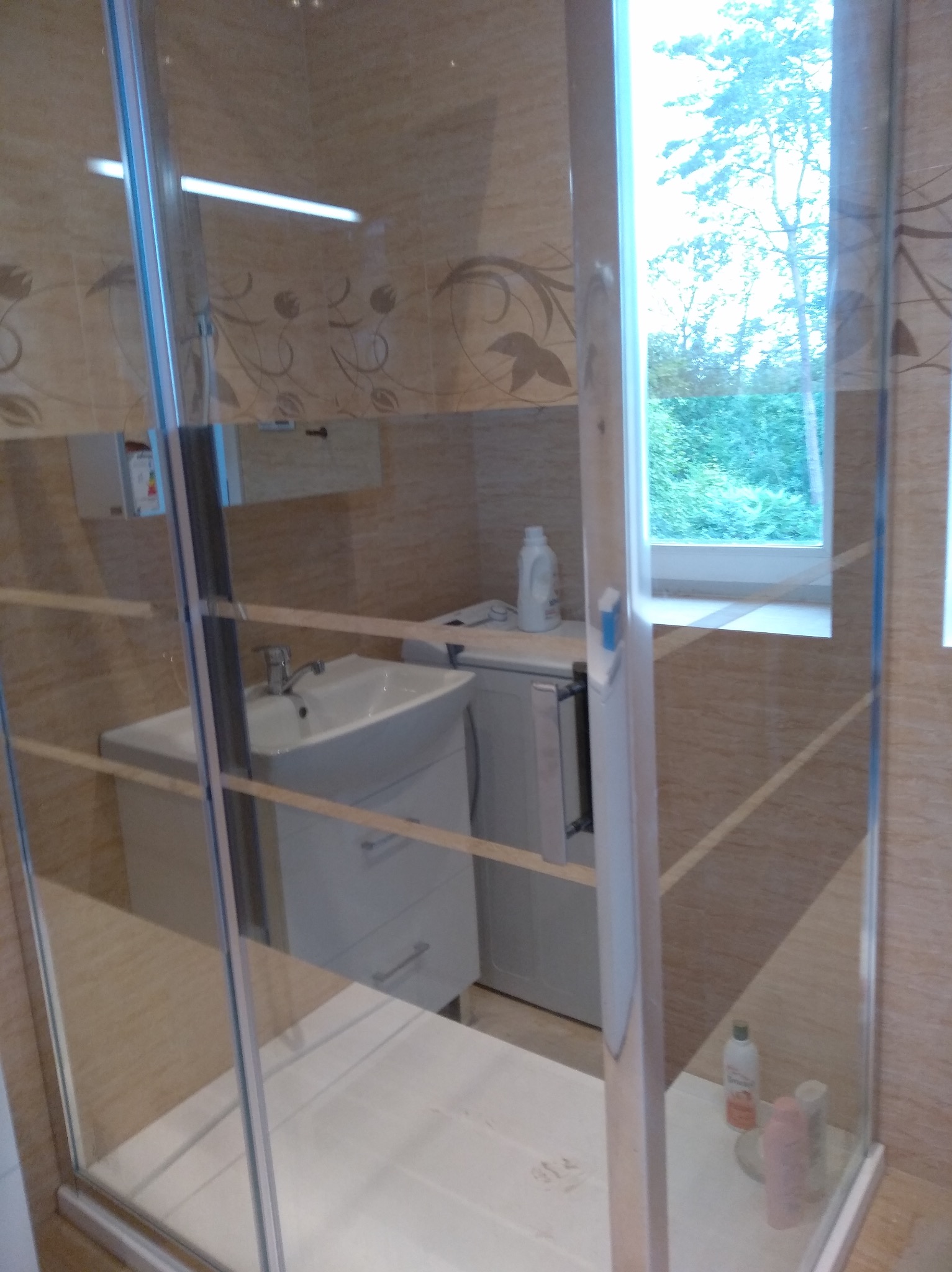 Wykończona łazienka z kabiną prysznicową typu walk-in, umywalką z szafką, pralką pod blatem i oknem z widokiem na zieleń.
