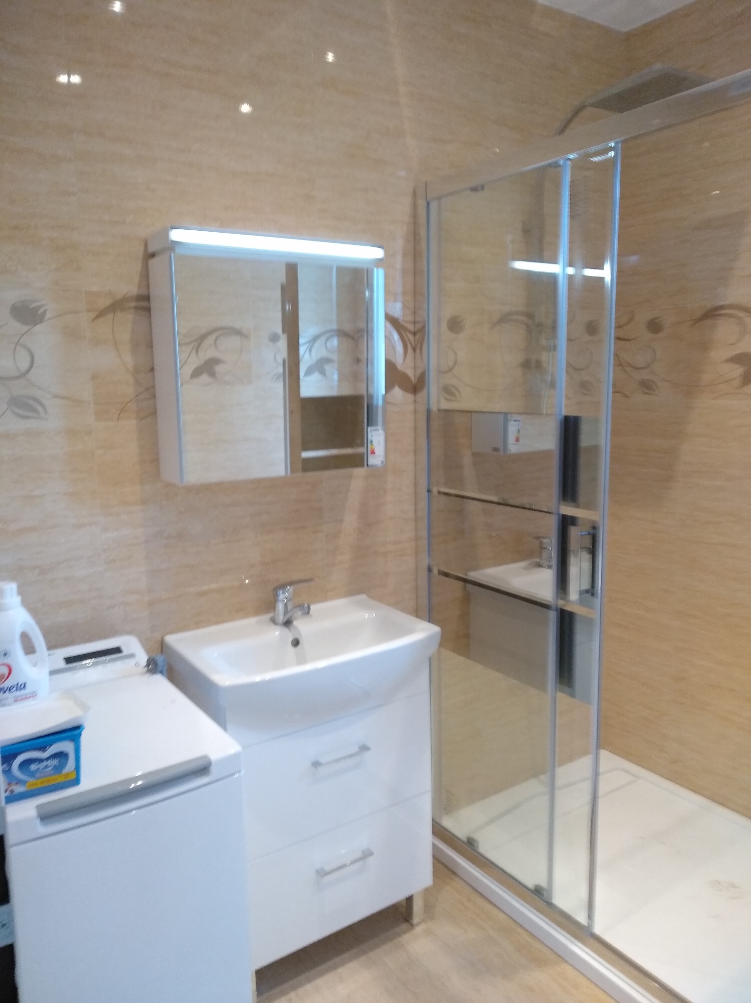 Wykończona łazienka z beżowymi płytkami, kabiną prysznicową ze szklanymi drzwiami przesuwnymi, umywalką z szafką i pralką z widocznymi środkami czystości.