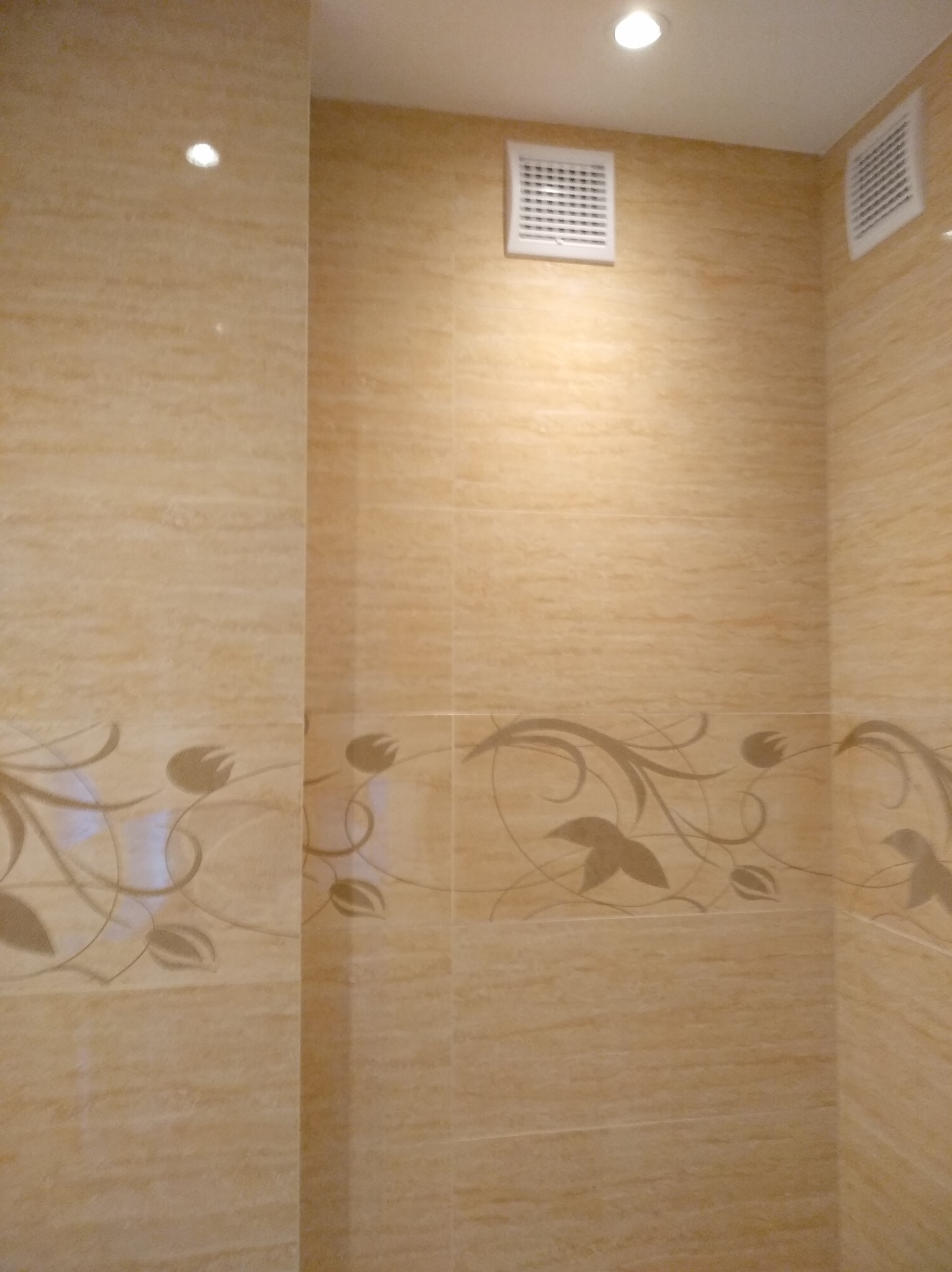 Wykończona łazienka z beżowymi płytkami imitującymi trawertyn, ozdobnymi motywami roślinnymi i kratkami wentylacyjnymi przy suficie.