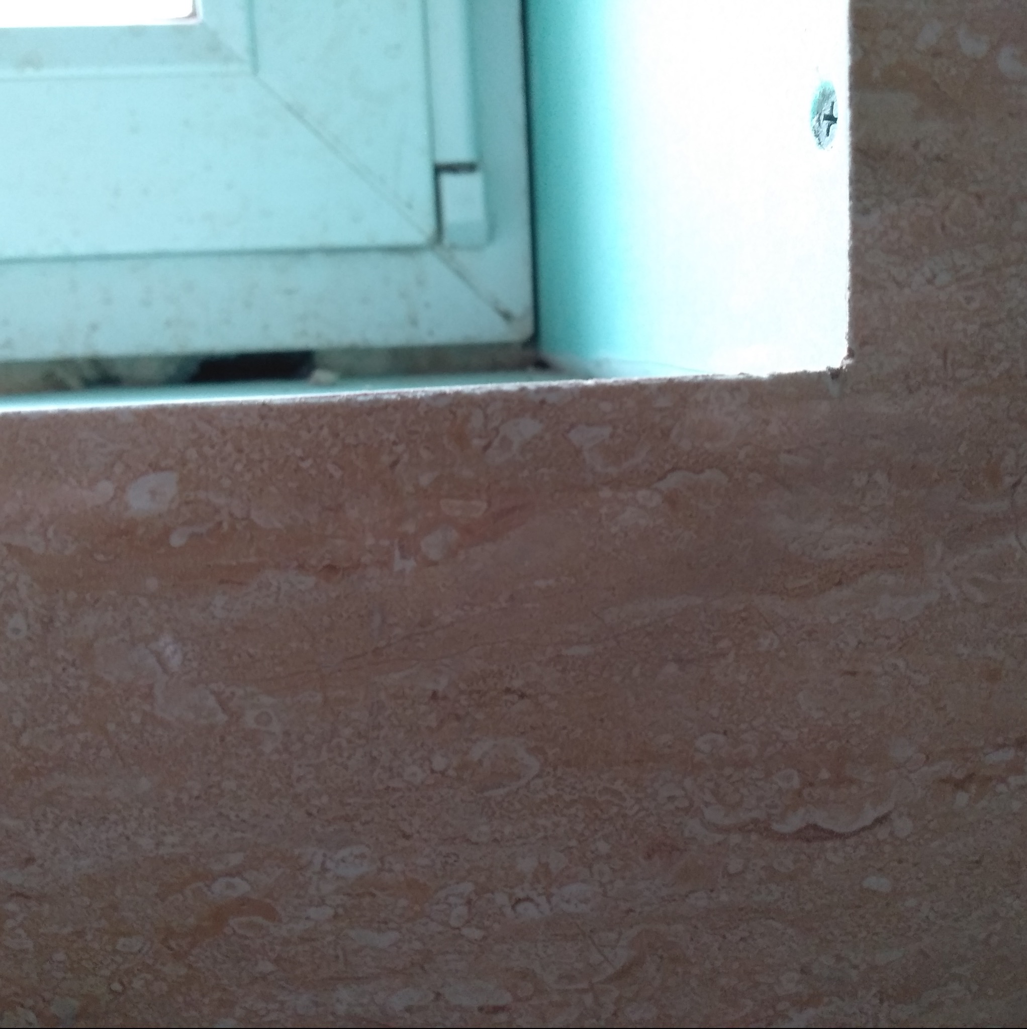 Fragment parapetu z beżowego marmuru, przylegający do ramy okiennej z widoczną śrubą mocującą.