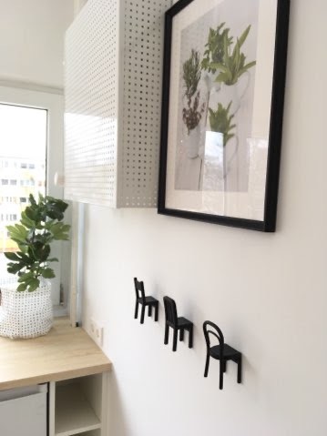 Jasne wnętrze z wiszącą szafką z perforowanej blachy, oprawionym obrazem z motywem roślinnym i trzema małymi, czarnymi wieszakami w kształcie krzeseł na białej ścianie, obok blatu z rośliną...