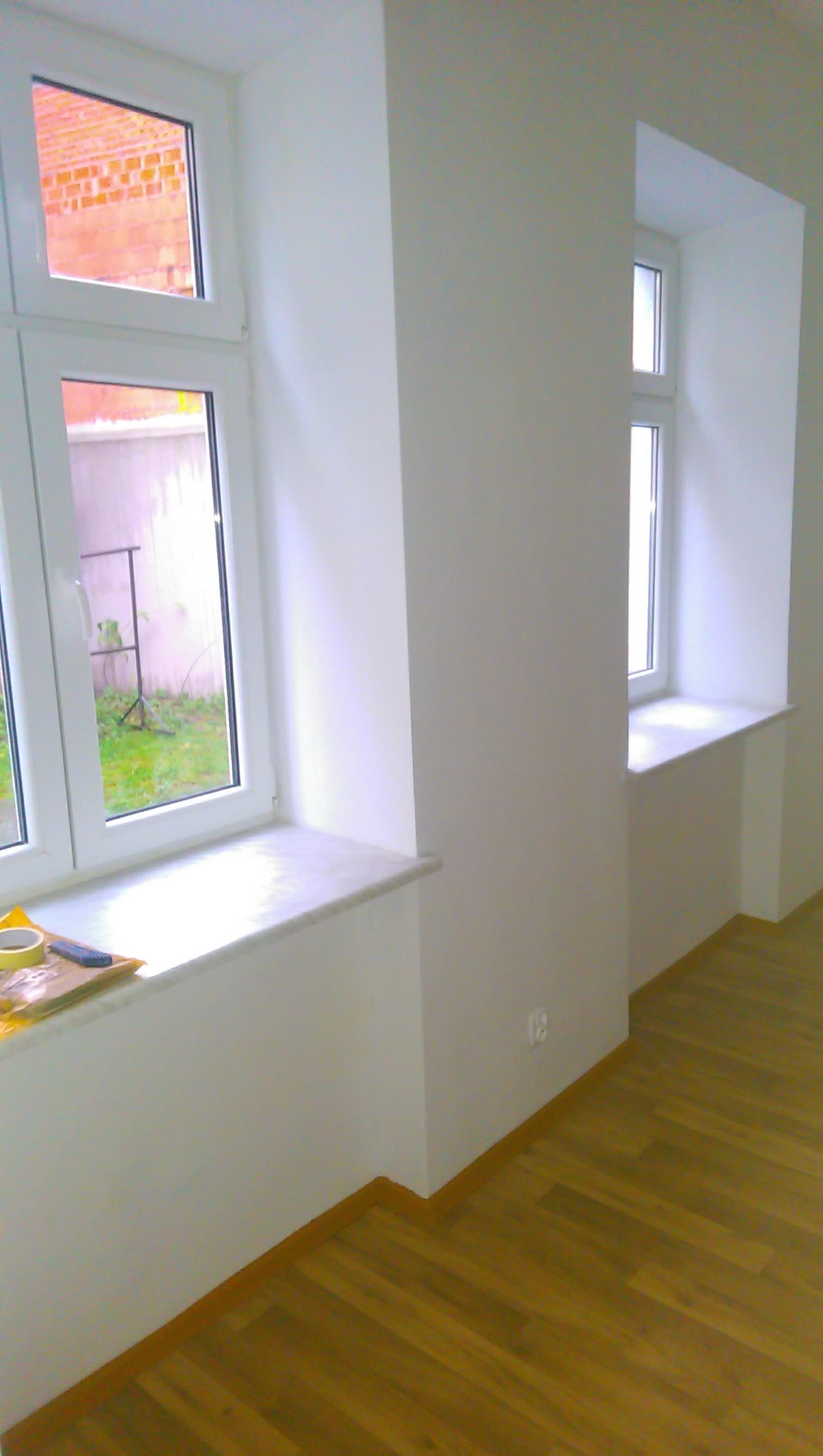 Białe, świeżo pomalowane ściany z dwoma oknami, marmurowe parapety, drewniana podłoga, widoczne ślady taśmy malarskiej na parapecie.