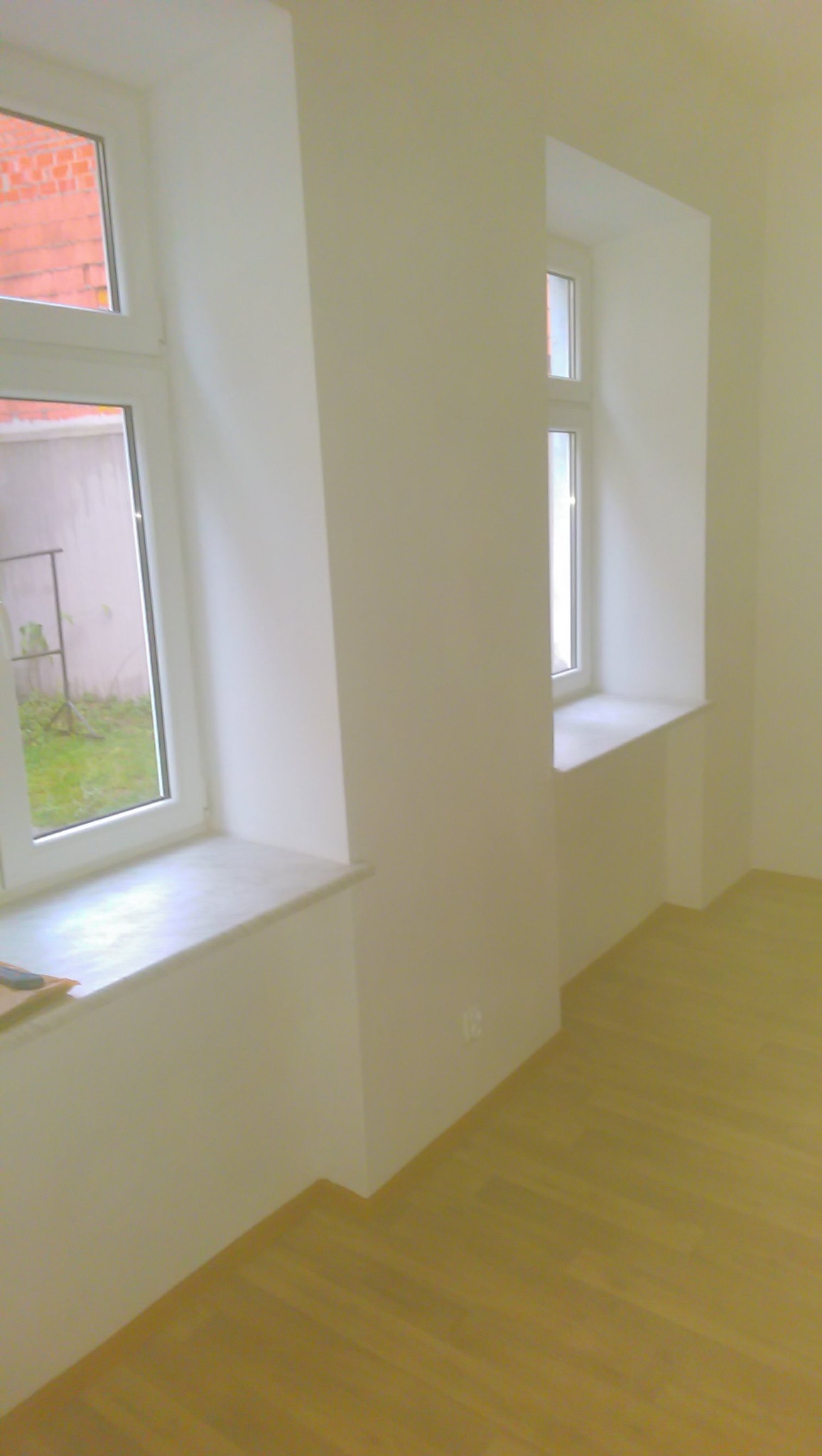 Pomalowane na biało wnętrze z dwoma oknami, jasną podłogą i gładkimi ścianami, widoczne gniazdko elektryczne.