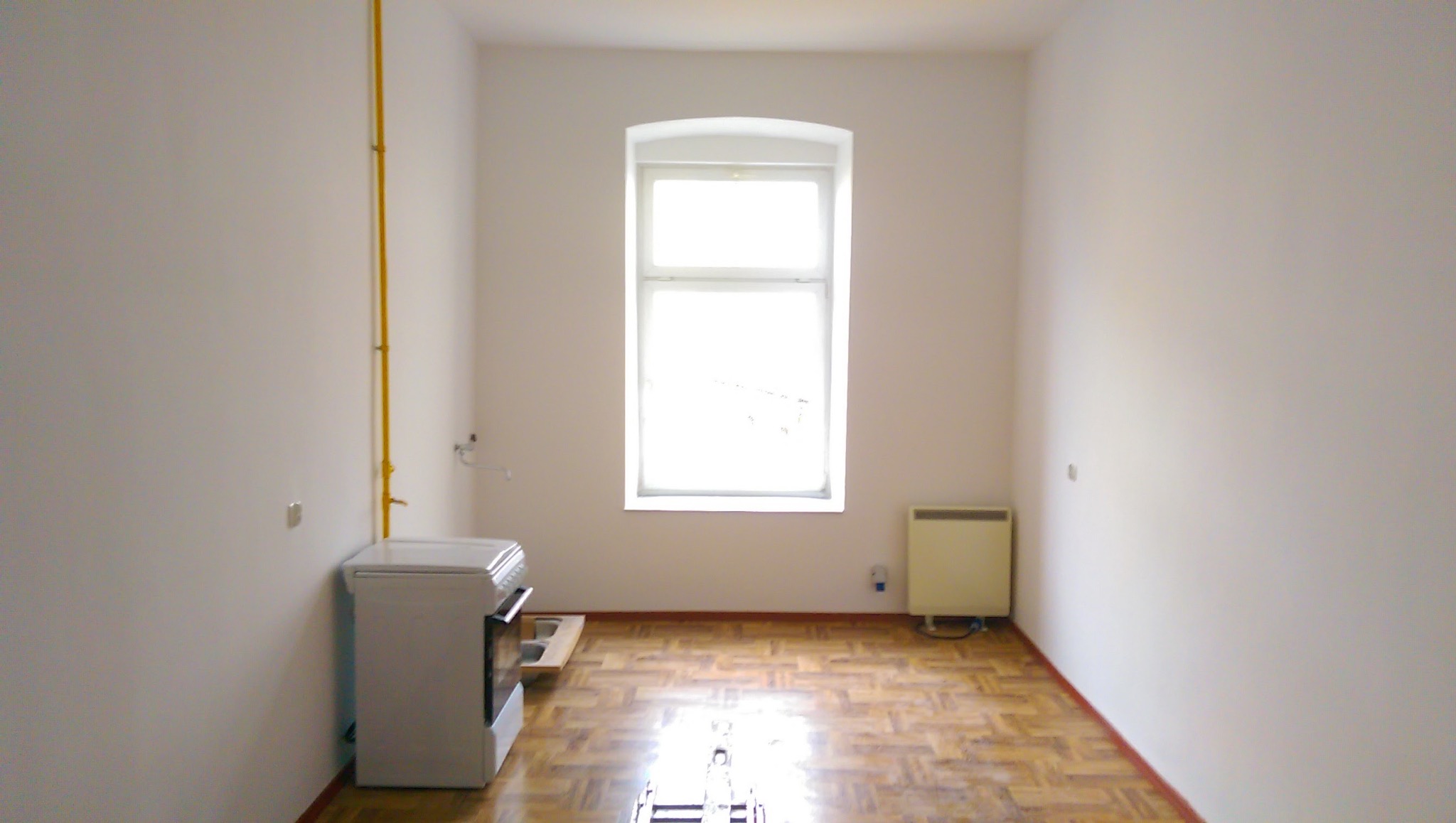 Pusty pokój po odświeżeniu z parkietem, świeżo pomalowanymi ścianami na biało, z żółtą rurą gazową przy ścianie, białą kuchenką i grzejnikiem elektrycznym pod oknem.