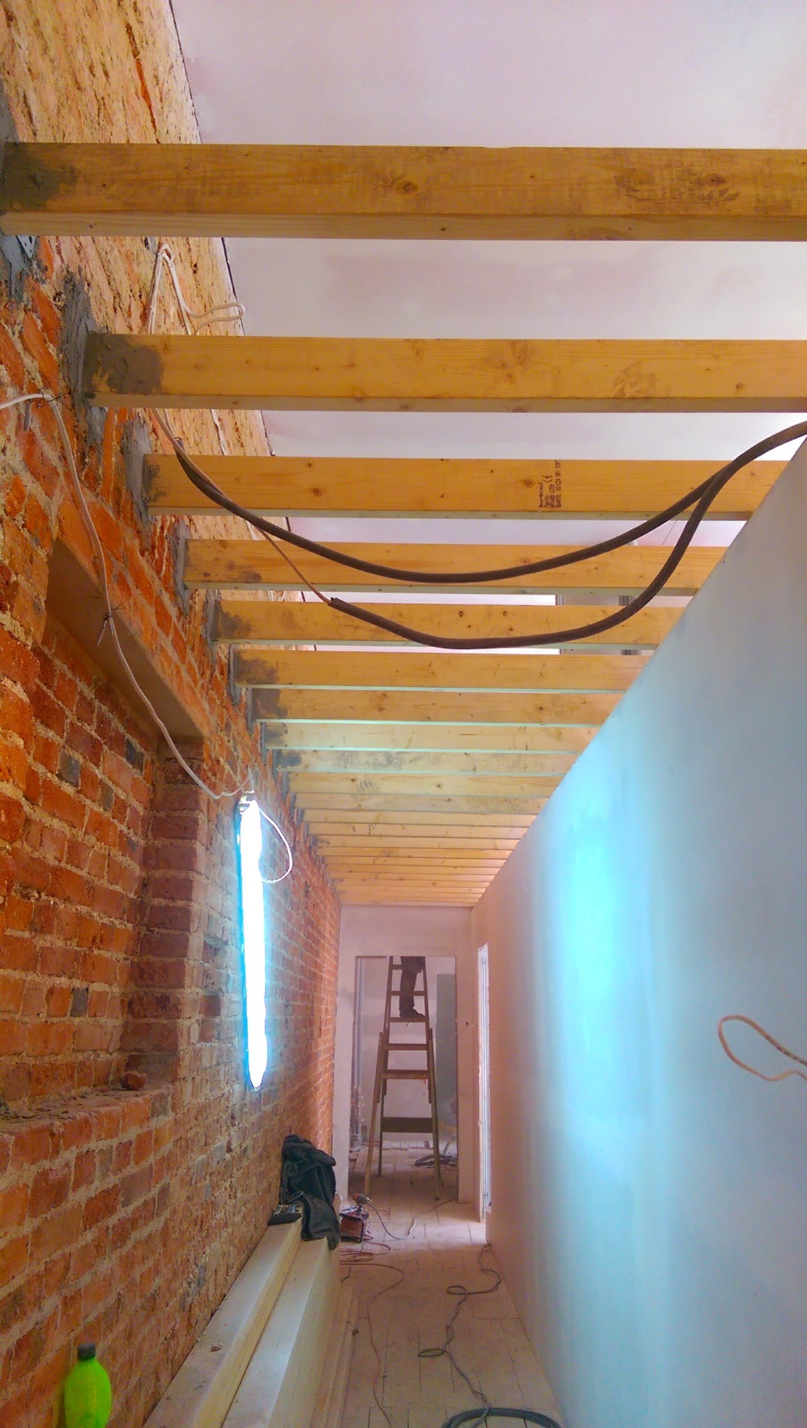 Wnętrze w trakcie prac: odsłonięta cegła, drewniana konstrukcja sufitu, instalacja elektryczna, drabina w głębi, widoczne etapy przygotowania do wykończenia.