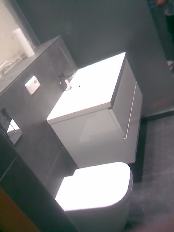 Nowoczesna toaleta podwieszana z białą szafką w łazience o szarych ścianach i podłodze, widoczny biały włącznik światła.