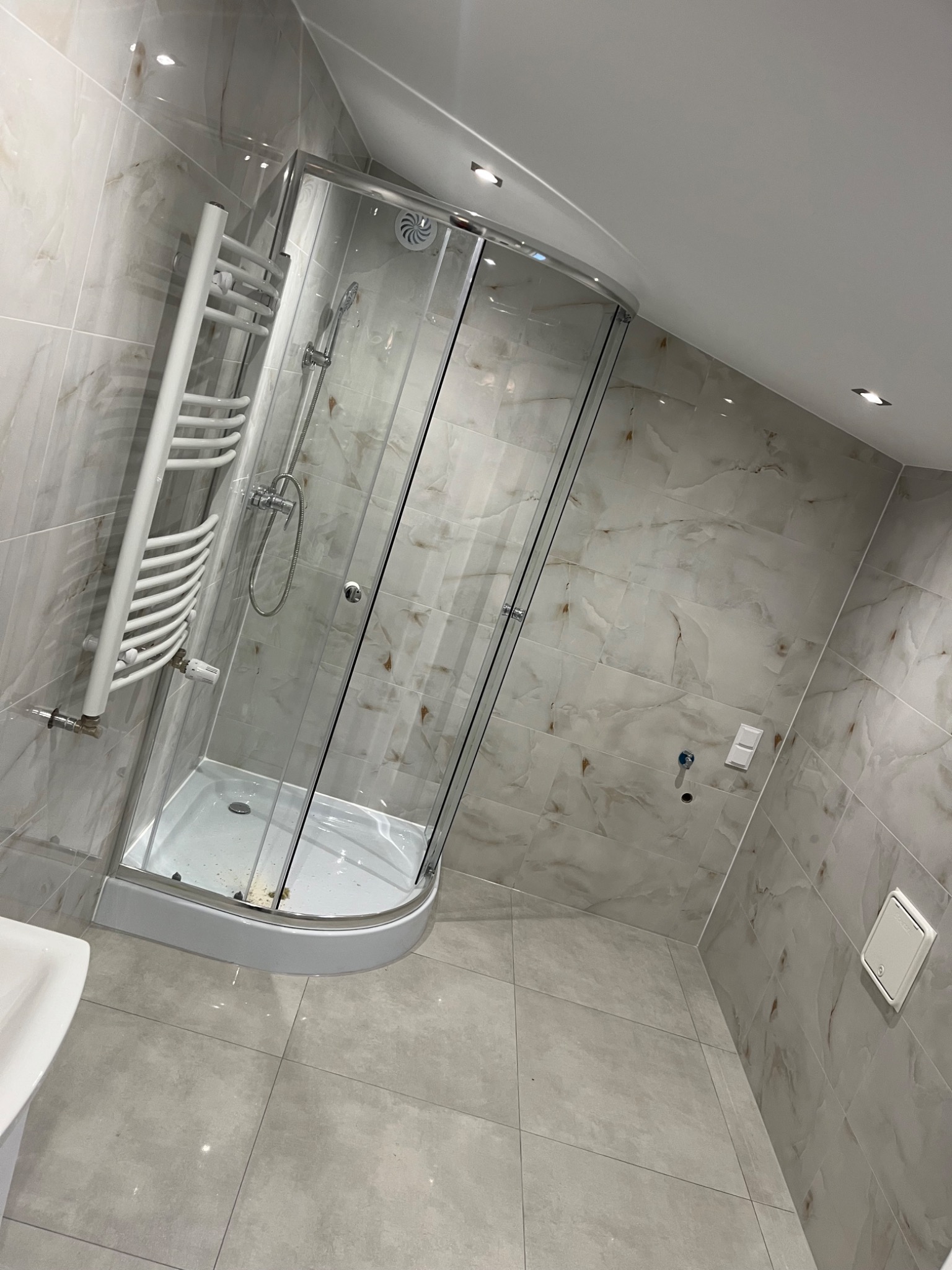 Wykończona łazienka z kabiną prysznicową, grzejnikiem drabinkowym i marmurowymi płytkami na ścianach. Nowoczesne oświetlenie punktowe.