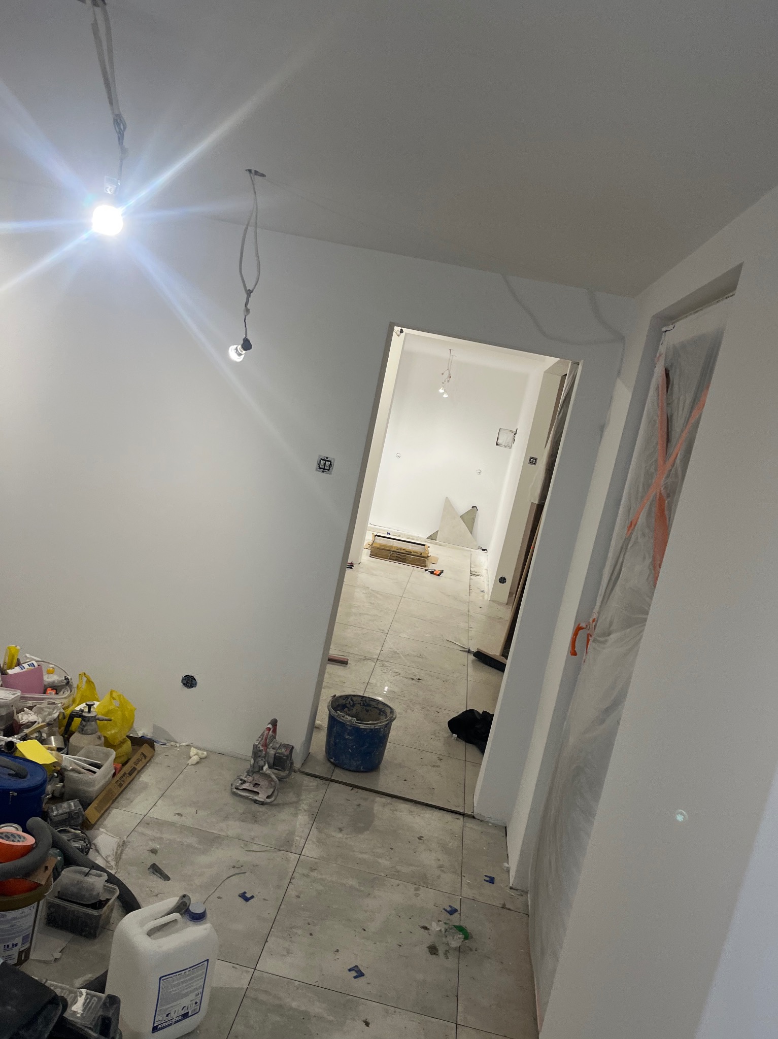 Wnętrze w trakcie prac wykończeniowych: świeżo pomalowane ściany, ułożona podłoga z płytek, widoczne narzędzia i materiały budowlane, surowe oświetlenie.