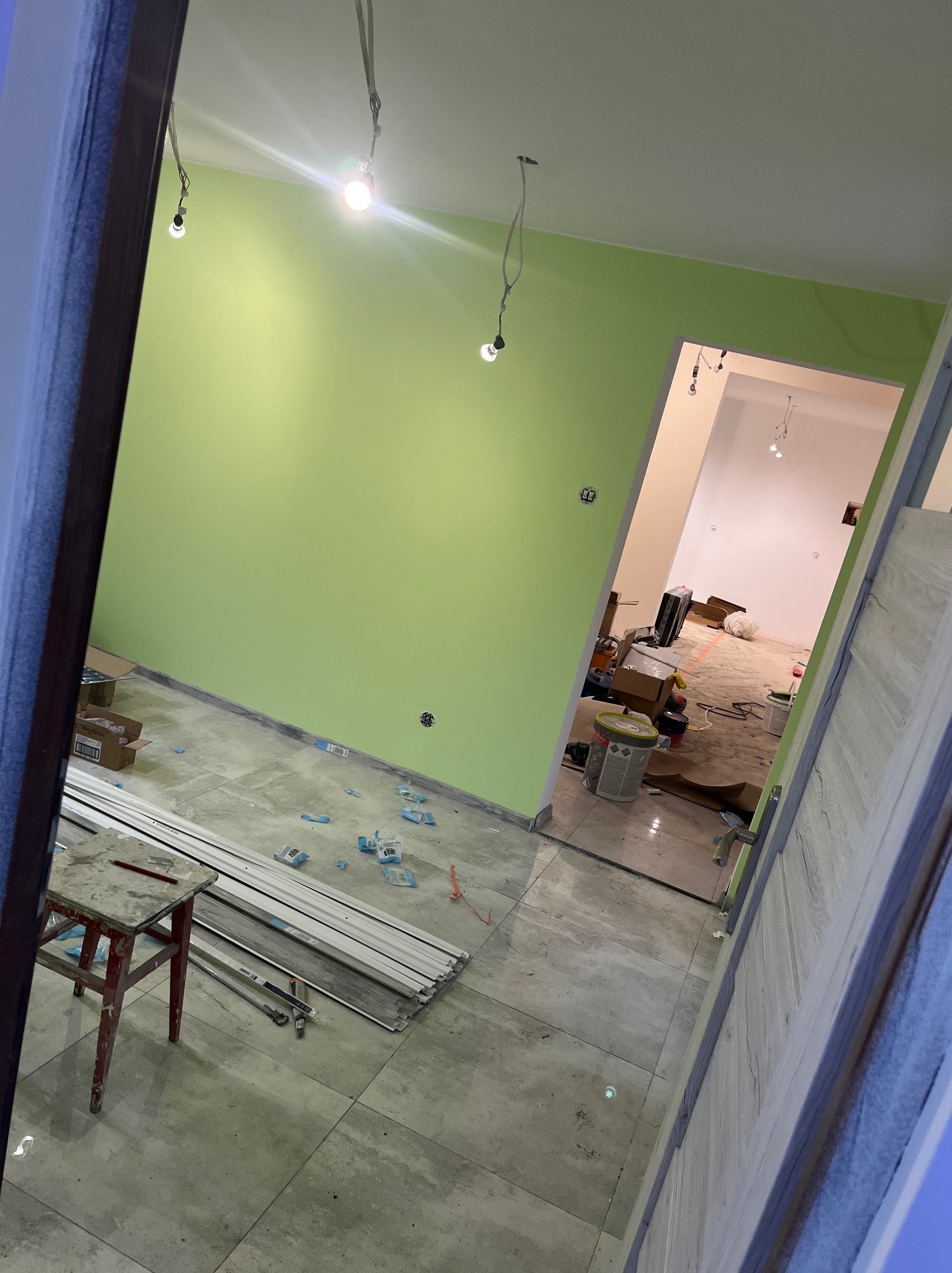 Wnętrze w trakcie prac wykończeniowych: świeżo pomalowana na zielono ściana, podłoga z płytek, widoczne listwy przypodłogowe i narzędzia. W tle otwarte drzwi do kolejnego pomieszczenia.