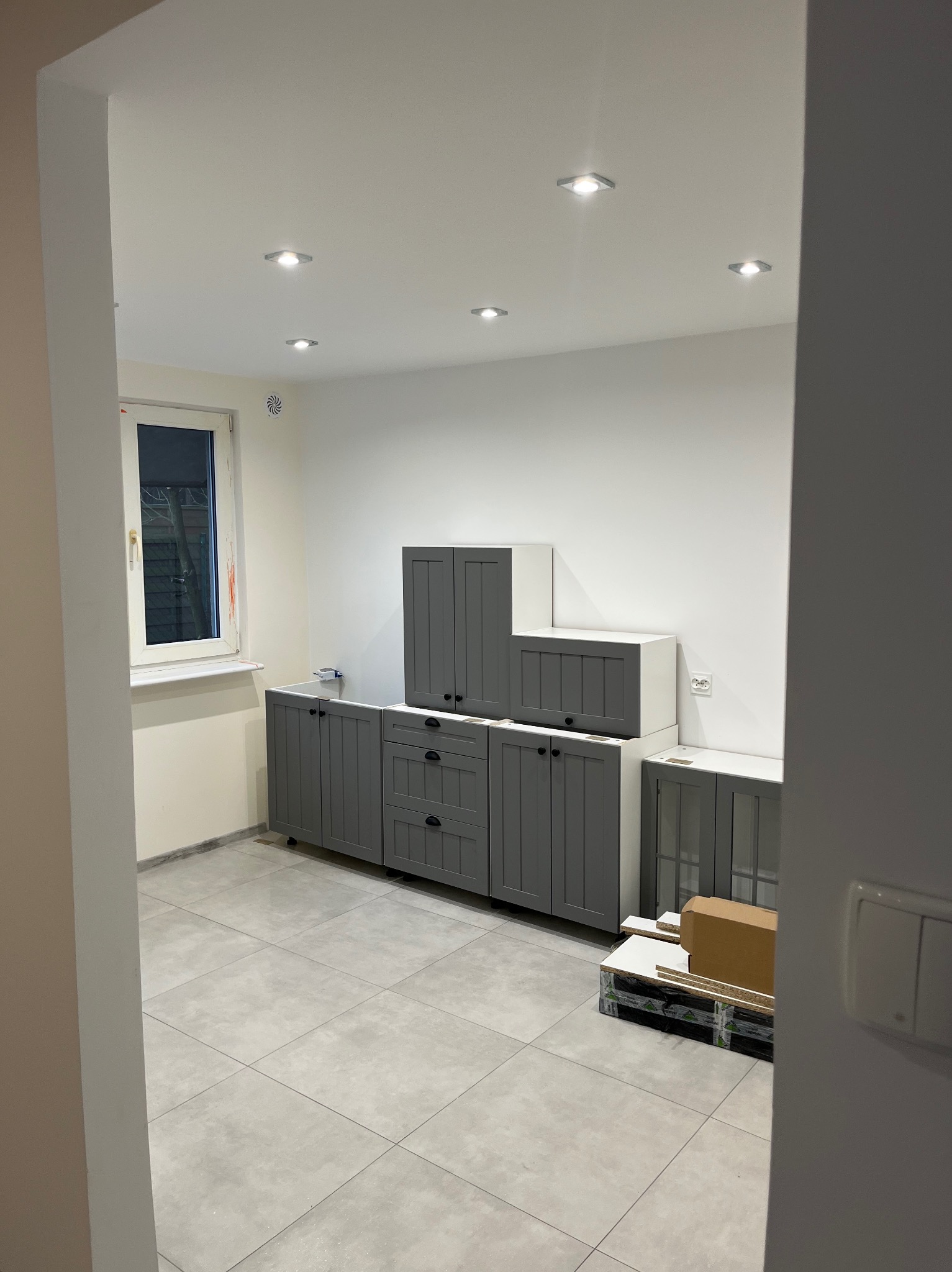Montaż szarych mebli kuchennych w Łodzi. Widok na częściowo zamontowaną zabudowę, białe ściany i szarą podłogę. Nowoczesne oświetlenie sufitowe.