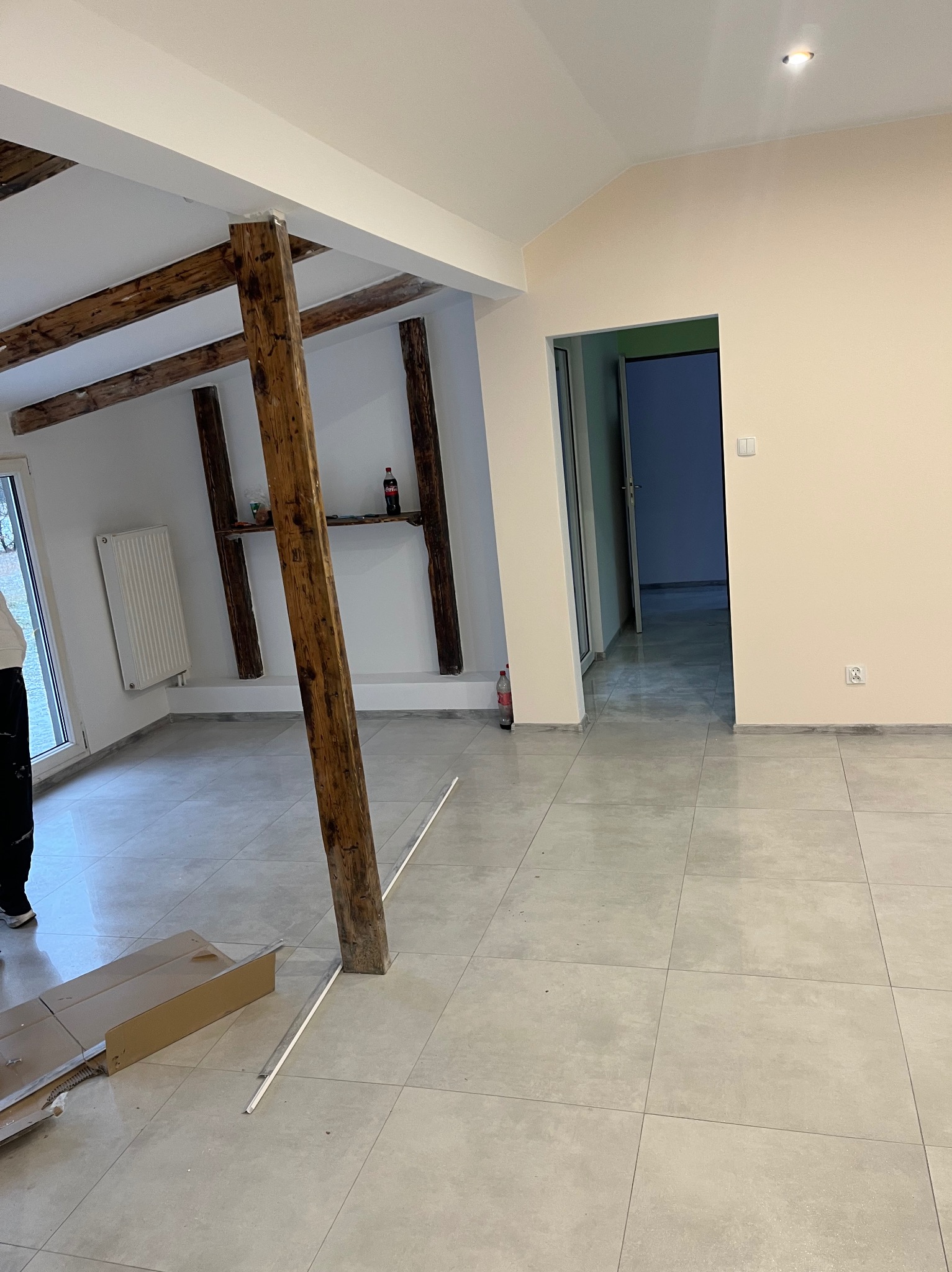 Jasne wnętrze po remoncie z drewnianymi belkami stropowymi i nową, jasną podłogą z płytek. Widoczne elementy wykończeniowe i fragment kartonu po panelach.