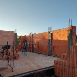 AWP-Domy Justyna Osowiecka - Wznoszenie ścian budynku z czerwonej ceramiki poryzowanej, widoczne szalunki słupów żelbetowych, rusztowania i stosy pustaków na placu budowy, w tle zieleń drzew.